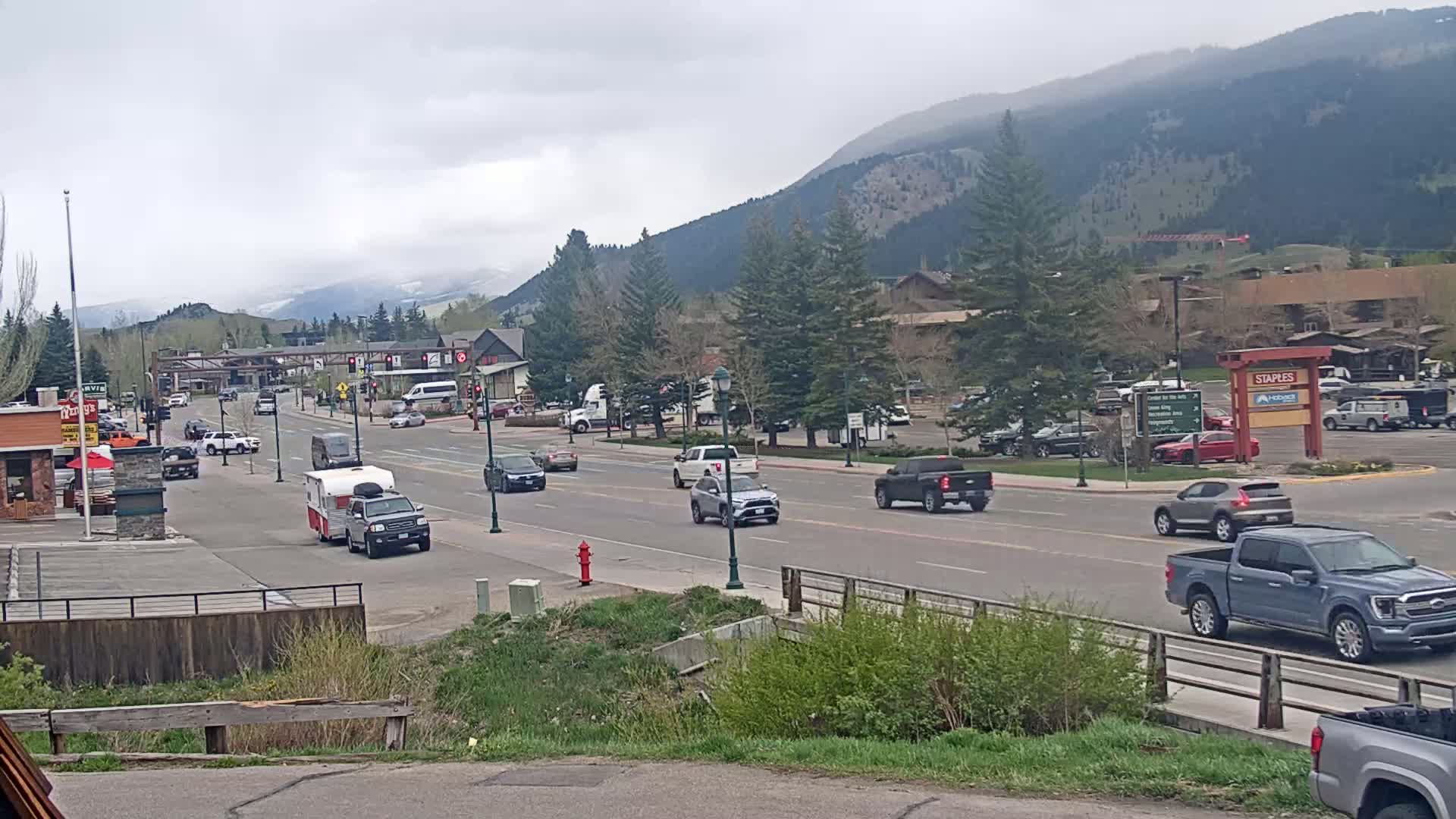 A partly cloudy day reveals a mountain-backed town with a main road populated by various vehicles, including trucks and SUVs,  and a small camper.