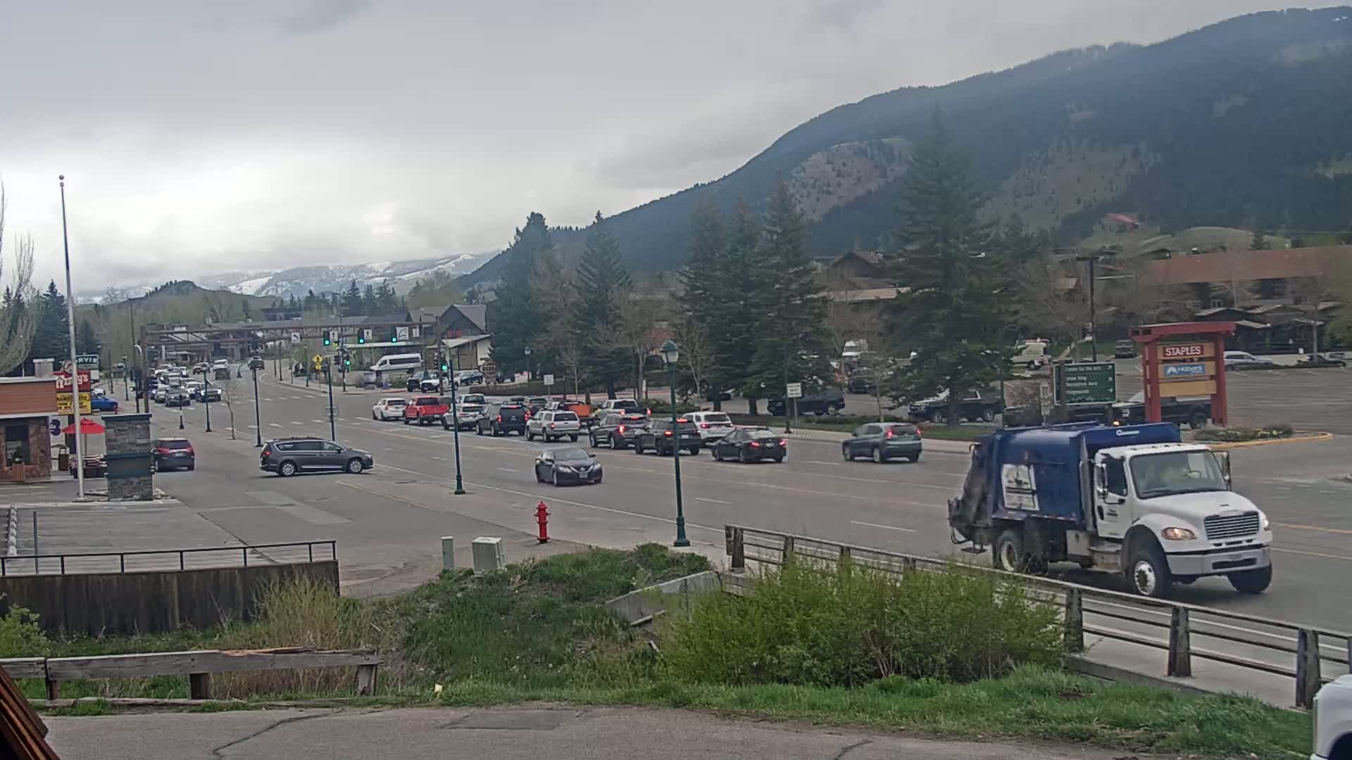 A line of cars drives along a road in a town nestled in a valley against a backdrop of mountains under an overcast sky.