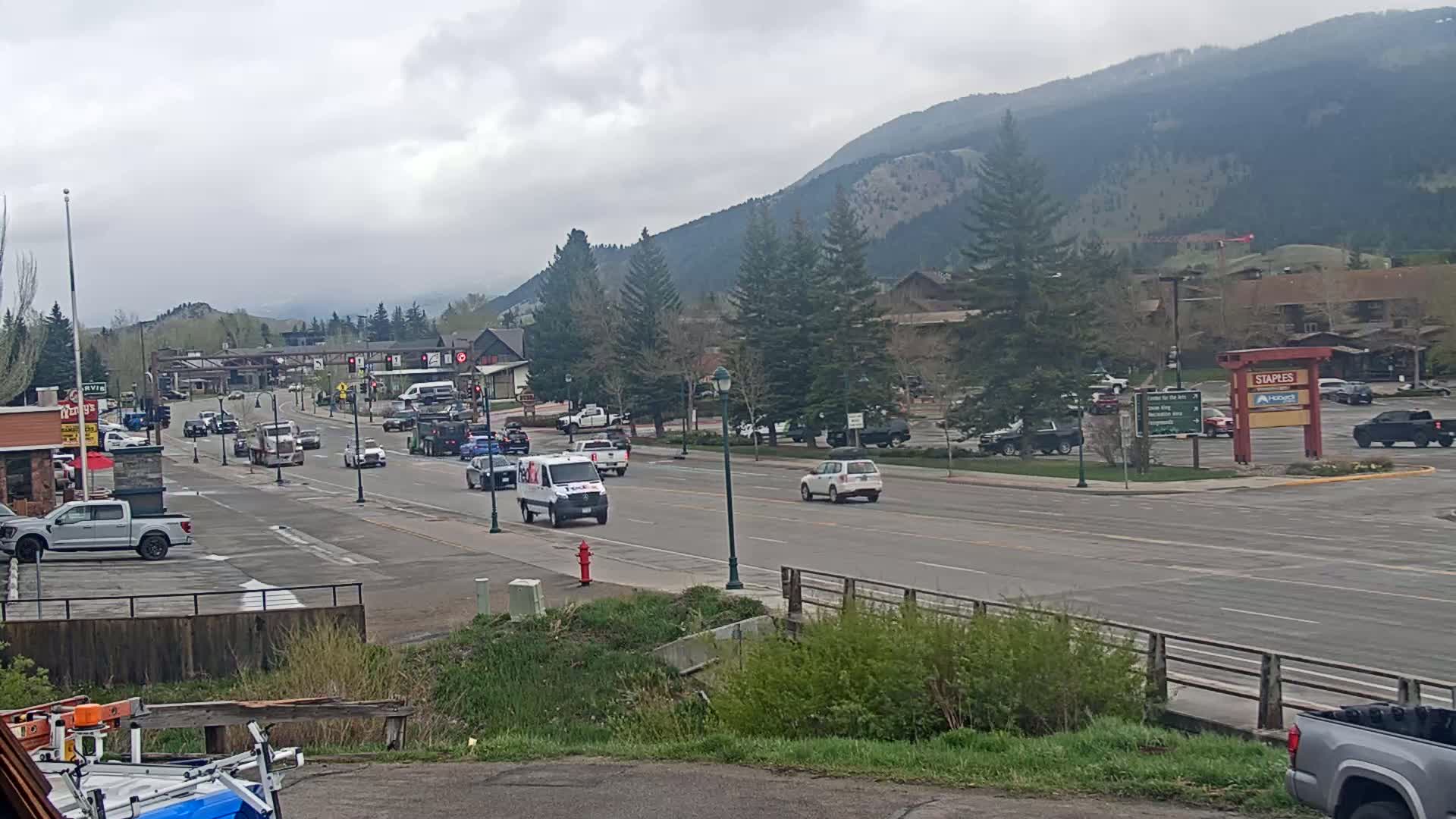 A street scene shows several vehicles traveling along a road that passes through a town nestled at the base of a mountain range under an overcast sky.