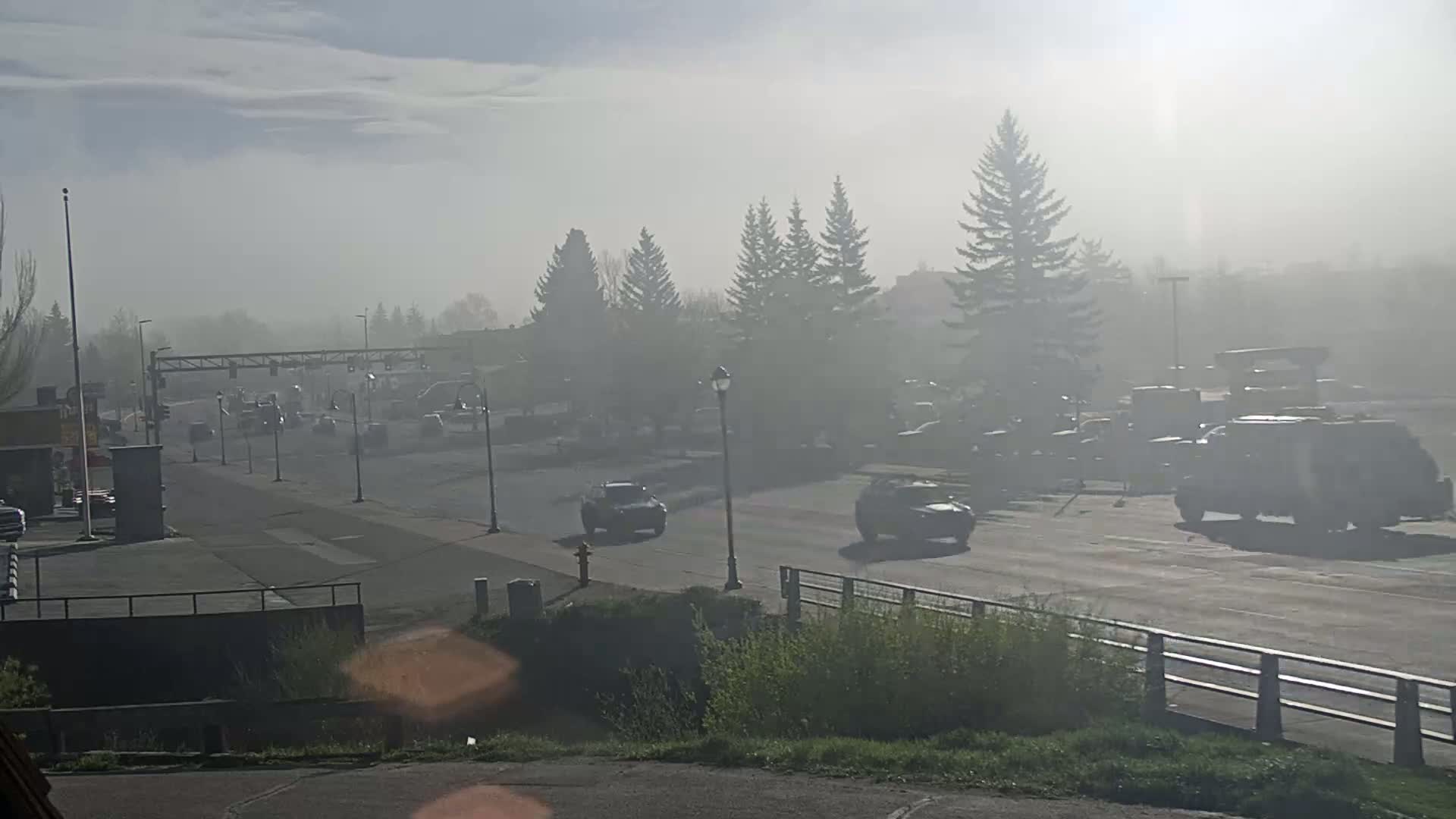 A hazy, foggy morning reveals a road with several vehicles and trees along a paved area, with a bridge partially visible in the foreground.