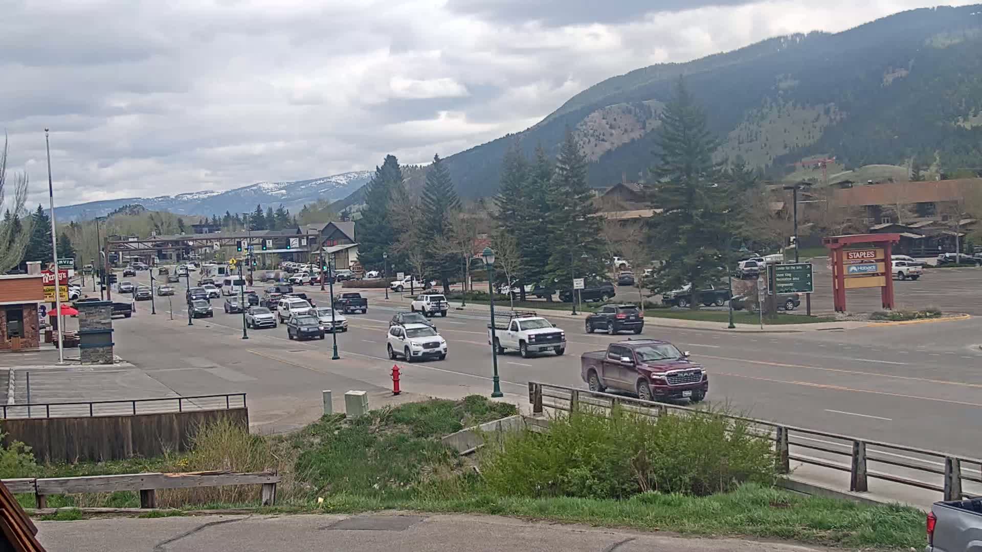 A mostly sunny day with some clouds shows a mountain town street with moderate traffic, lined with businesses and trees, with mountains in the background.