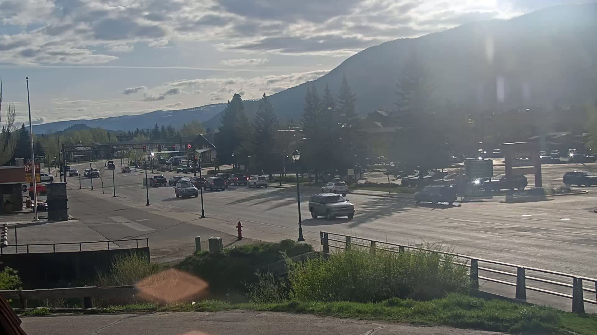A partly cloudy day shows a road with moderate traffic in front of buildings and trees, with mountains in the background.