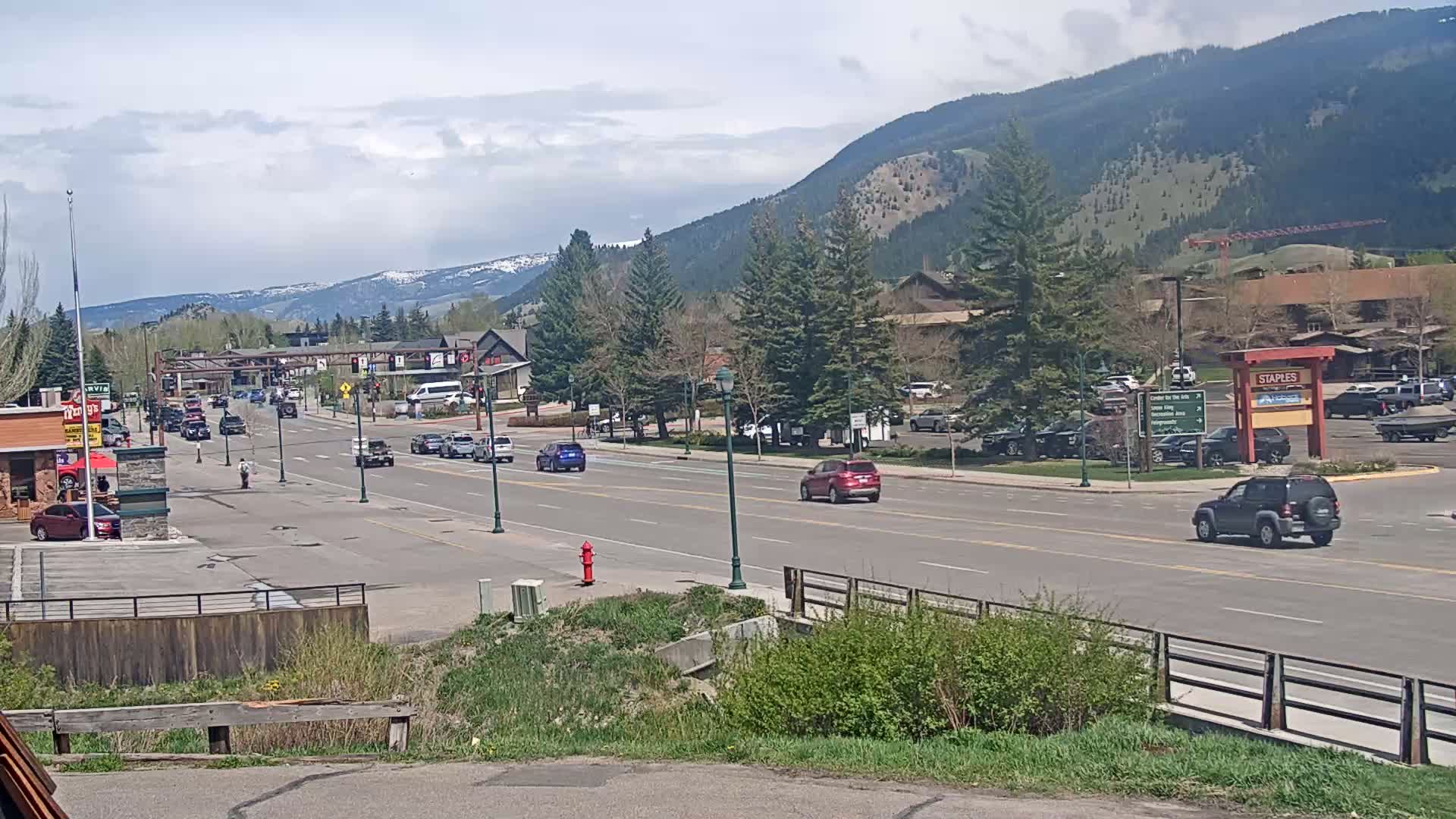 A partly cloudy day shows a town's main street with cars driving on a multi-lane road, lined with buildings and trees, nestled in a valley with mountains in the background.