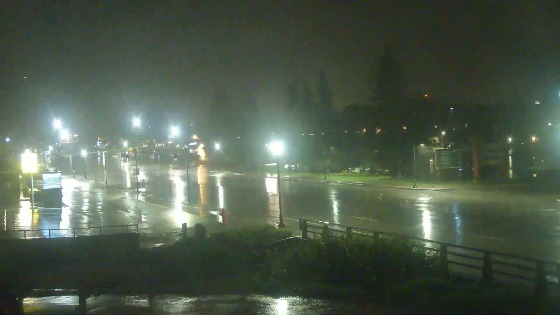 A rain-slicked, empty street at night is illuminated by streetlights.