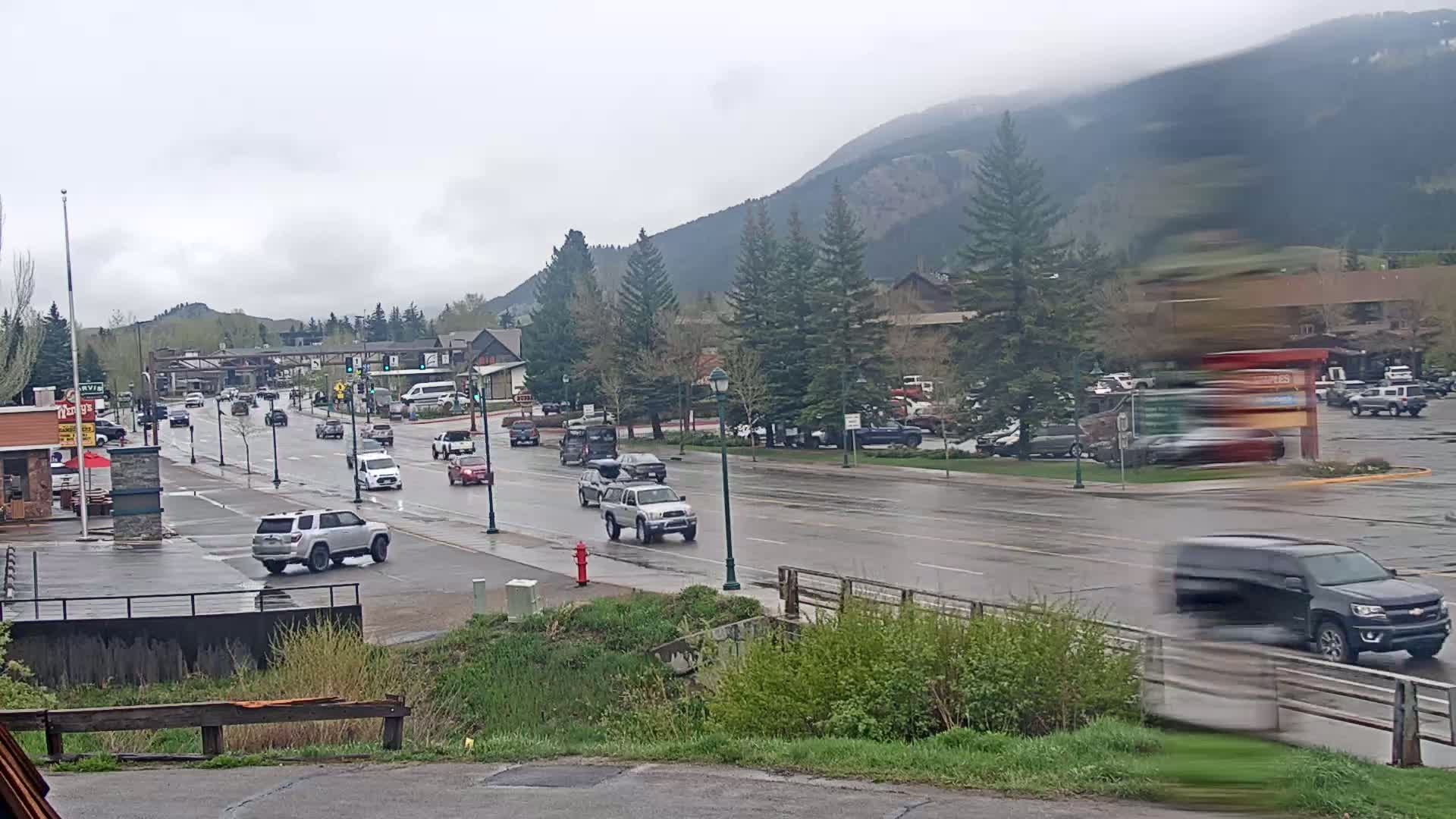 A wet, paved road with several cars driving through a town in front of a mountain range under an overcast sky.