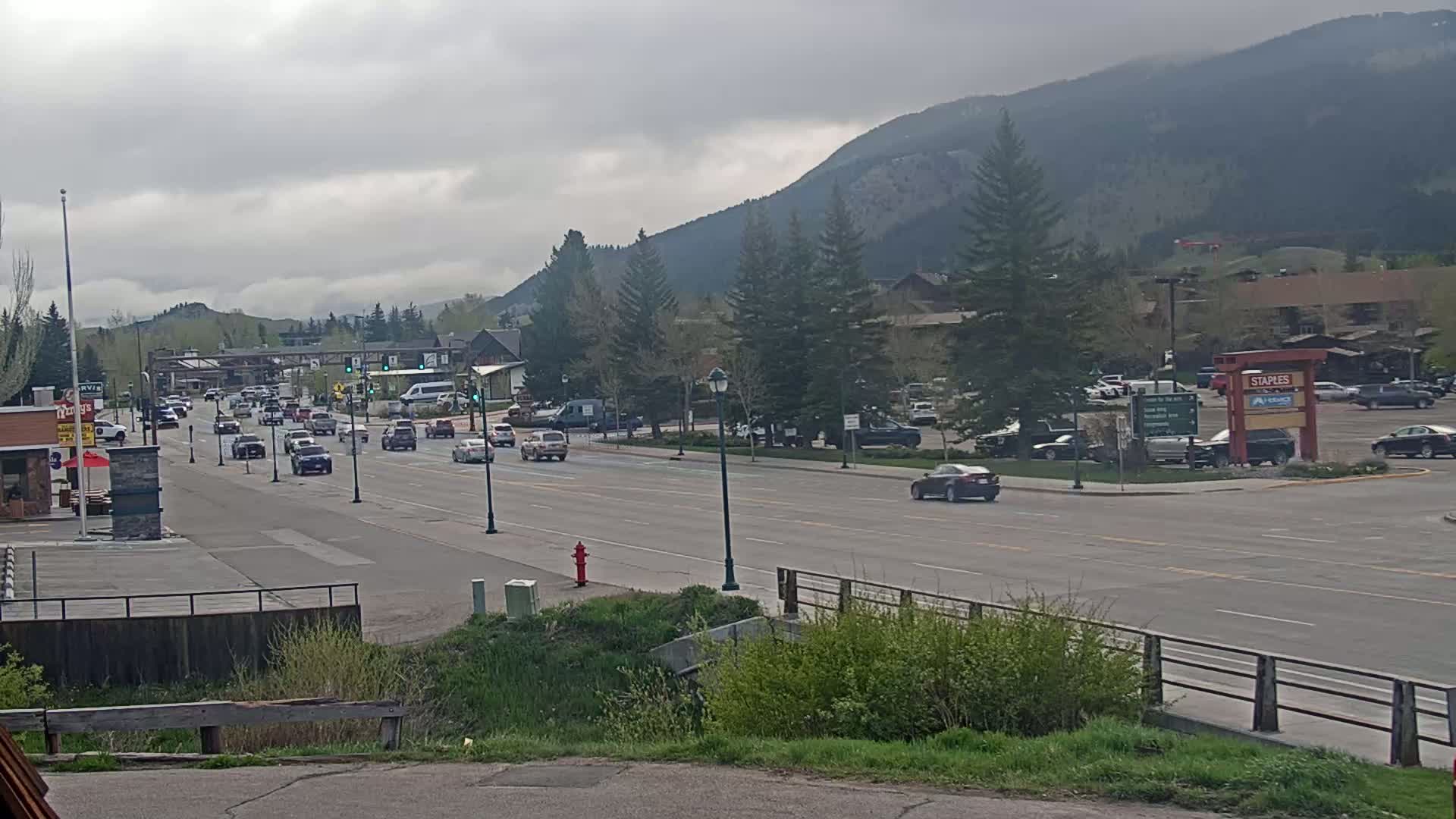 A partly cloudy day reveals a town street with moderate traffic, lined with trees and buildings, set against a backdrop of mountains.