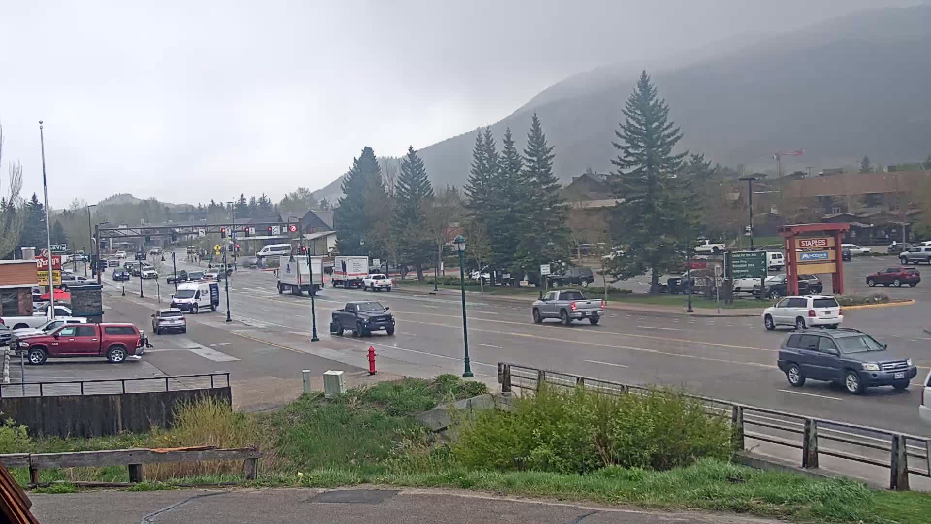 A wet, overcast day shows a multi-lane road with several vehicles driving through a town nestled at the base of a mountain, surrounded by evergreen trees.