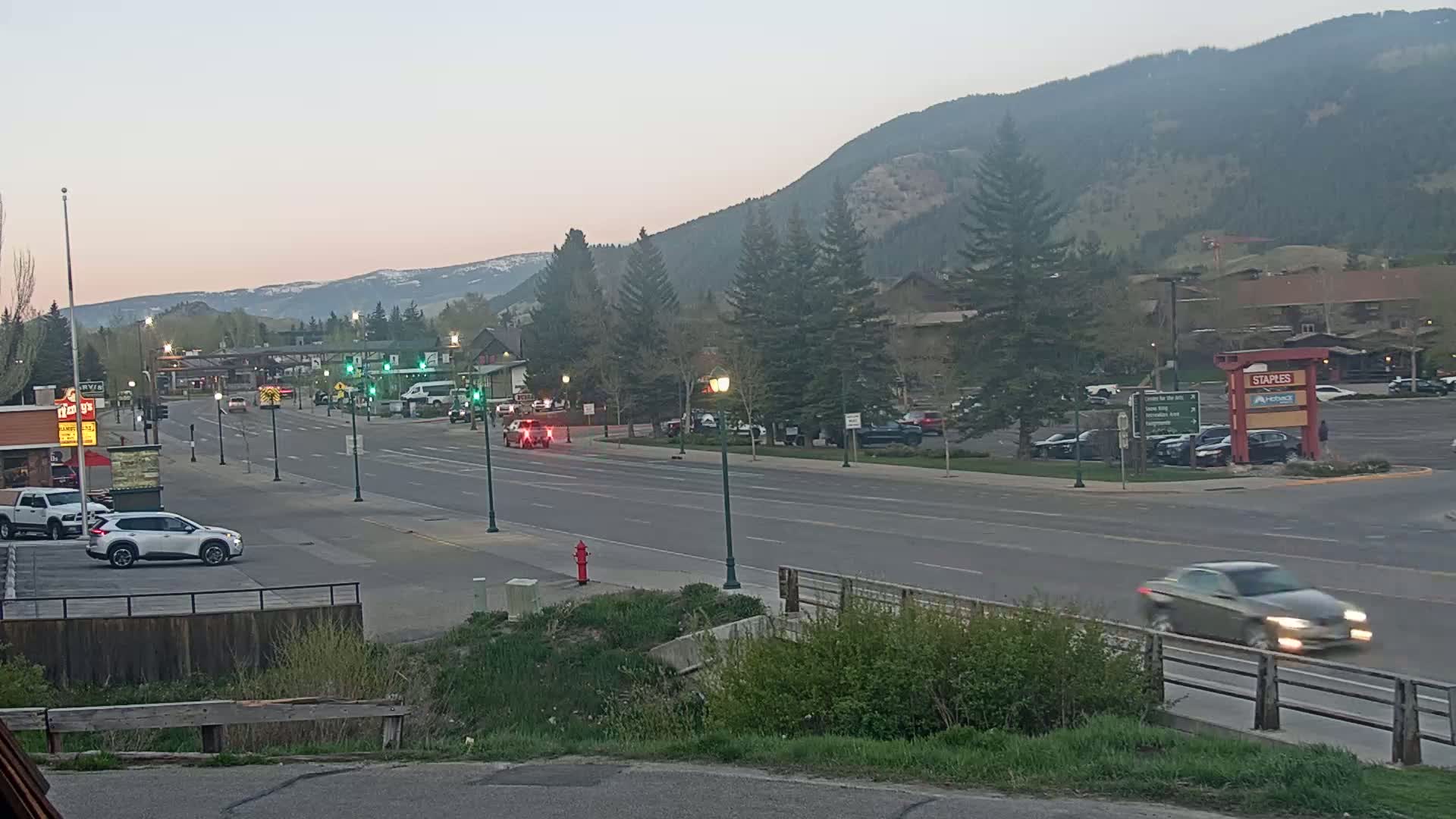 A twilight view of a multi-lane road in a town nestled at the base of a mountain range, with sparse traffic and several parked vehicles visible.