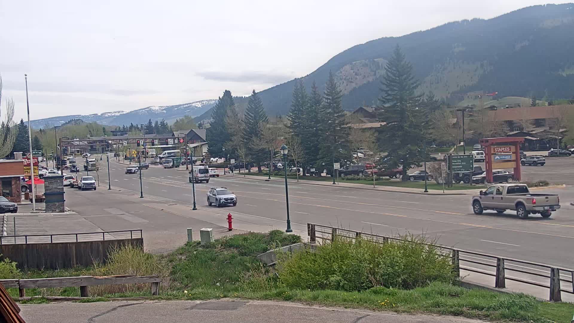 A partly cloudy day reveals a town street scene with cars driving on a multi-lane road lined with trees, businesses, and parking lots, set against a backdrop of mountains.