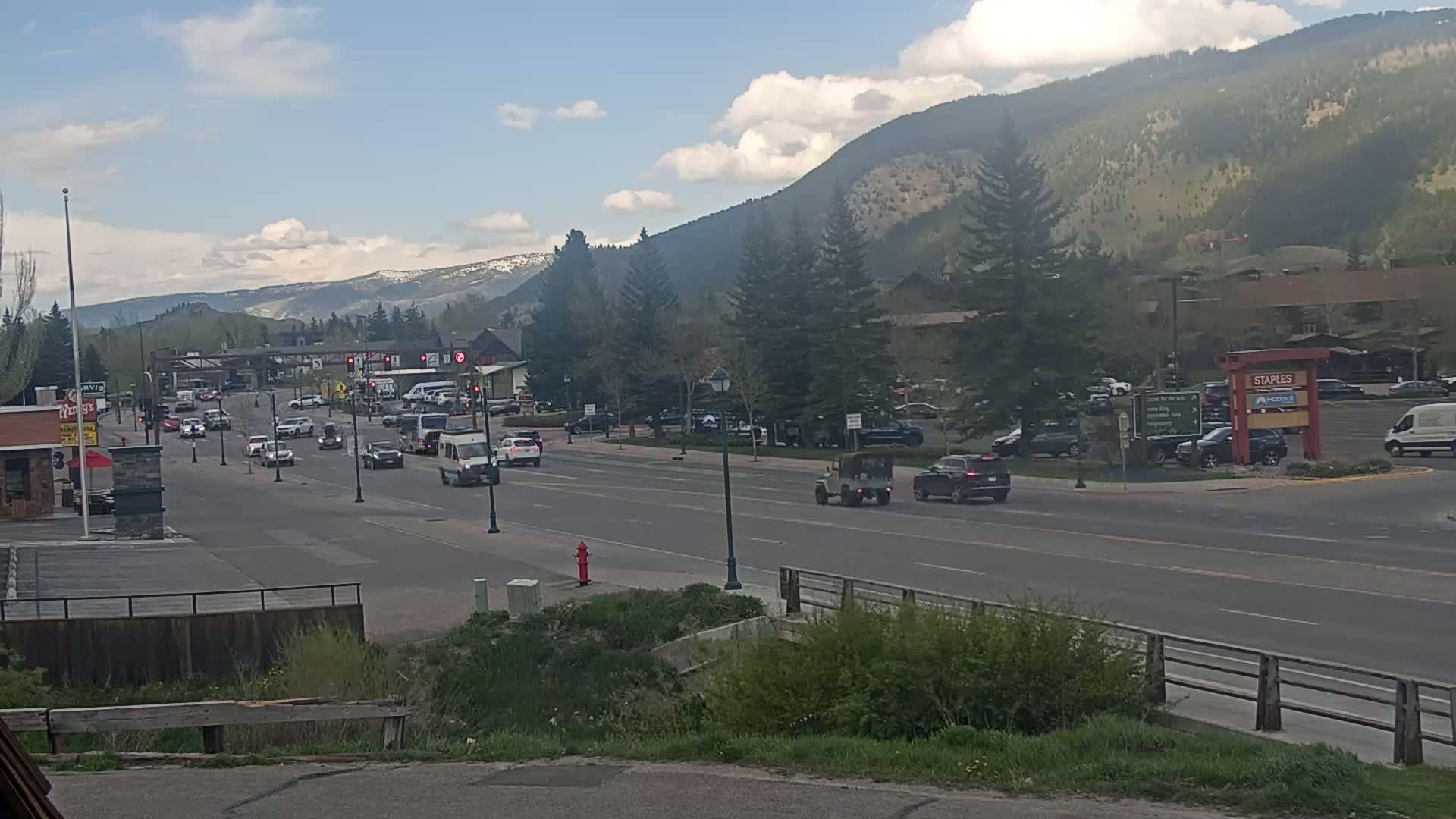 A partly cloudy day reveals a mountain range in the background of a multi-lane road with moderate traffic, lined with buildings and trees.