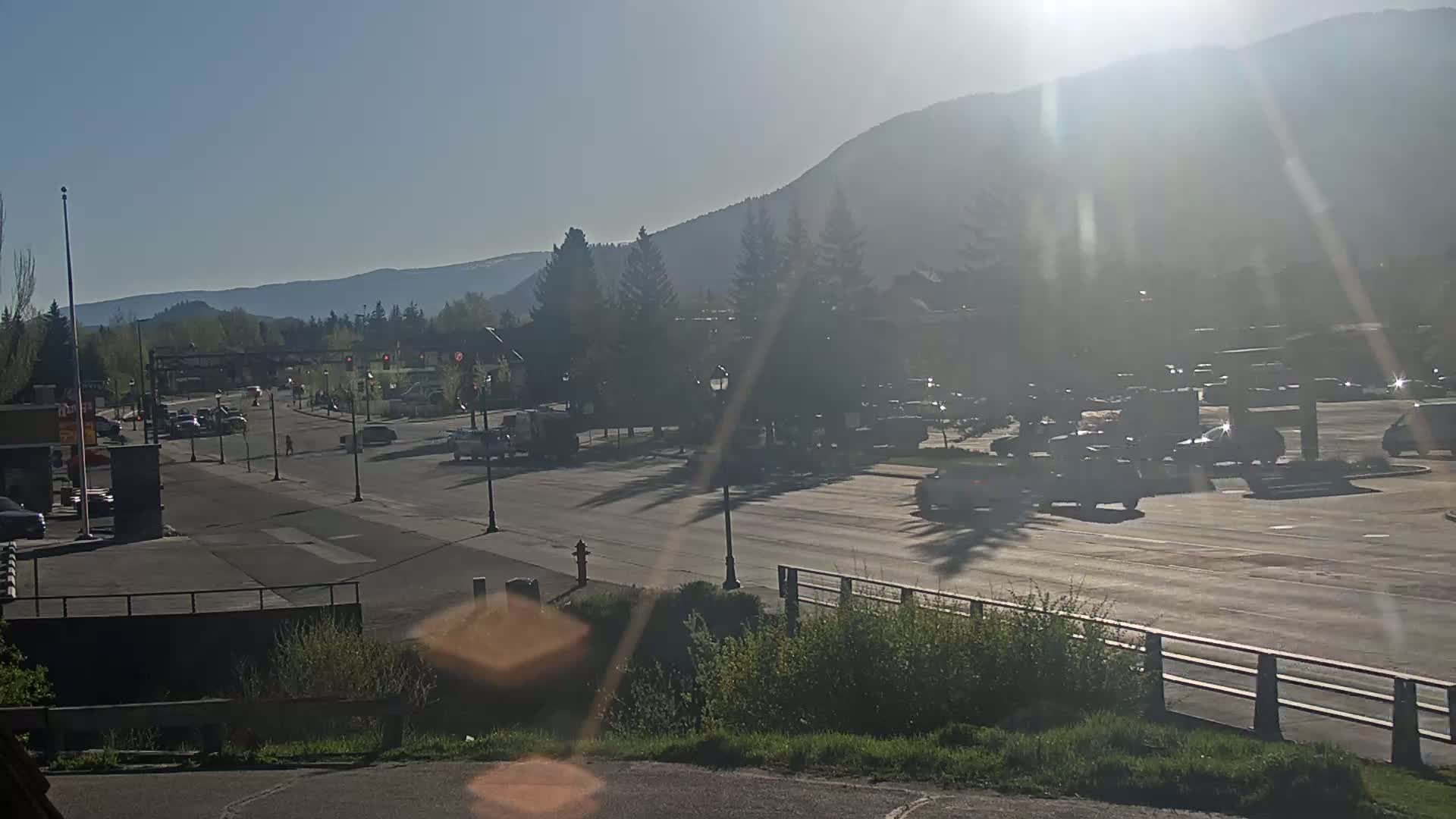 A sunny day reveals a roadway with cars parked and driving past buildings and trees, with mountains visible in the background.
