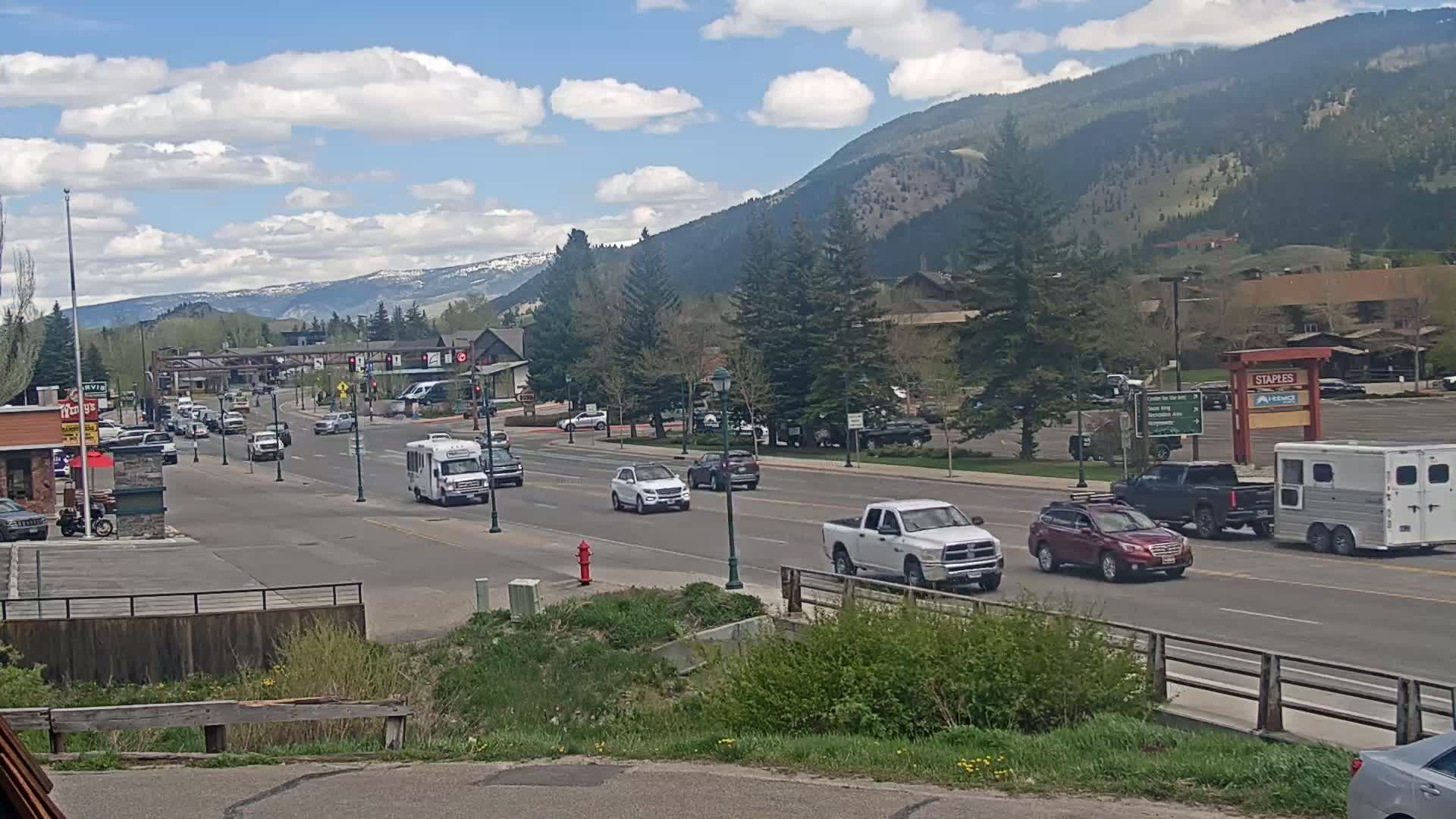 A partly cloudy day shows a mountain-backed town street with several cars, a van, and a horse trailer driving along a multi-lane road.