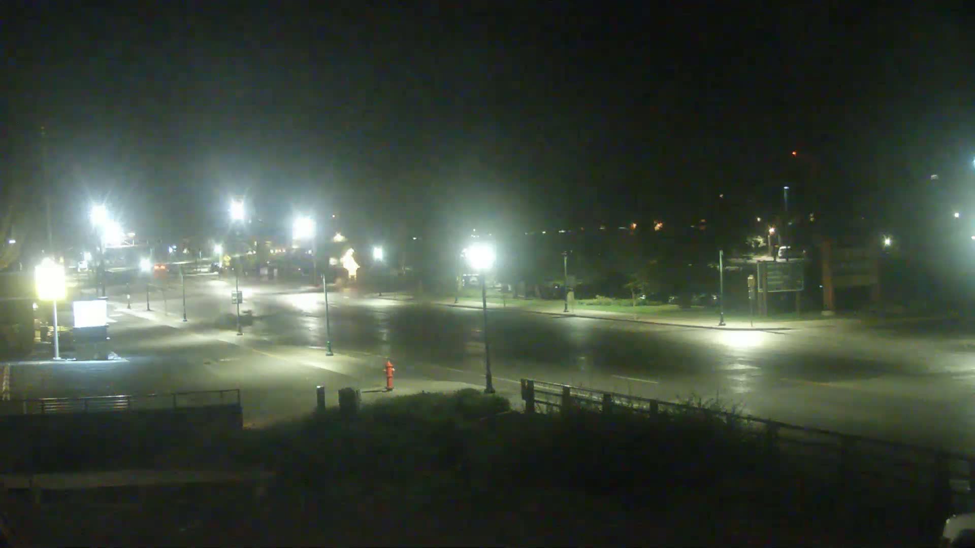 A wet, empty street at night is illuminated by streetlights, with a few distant vehicles visible.