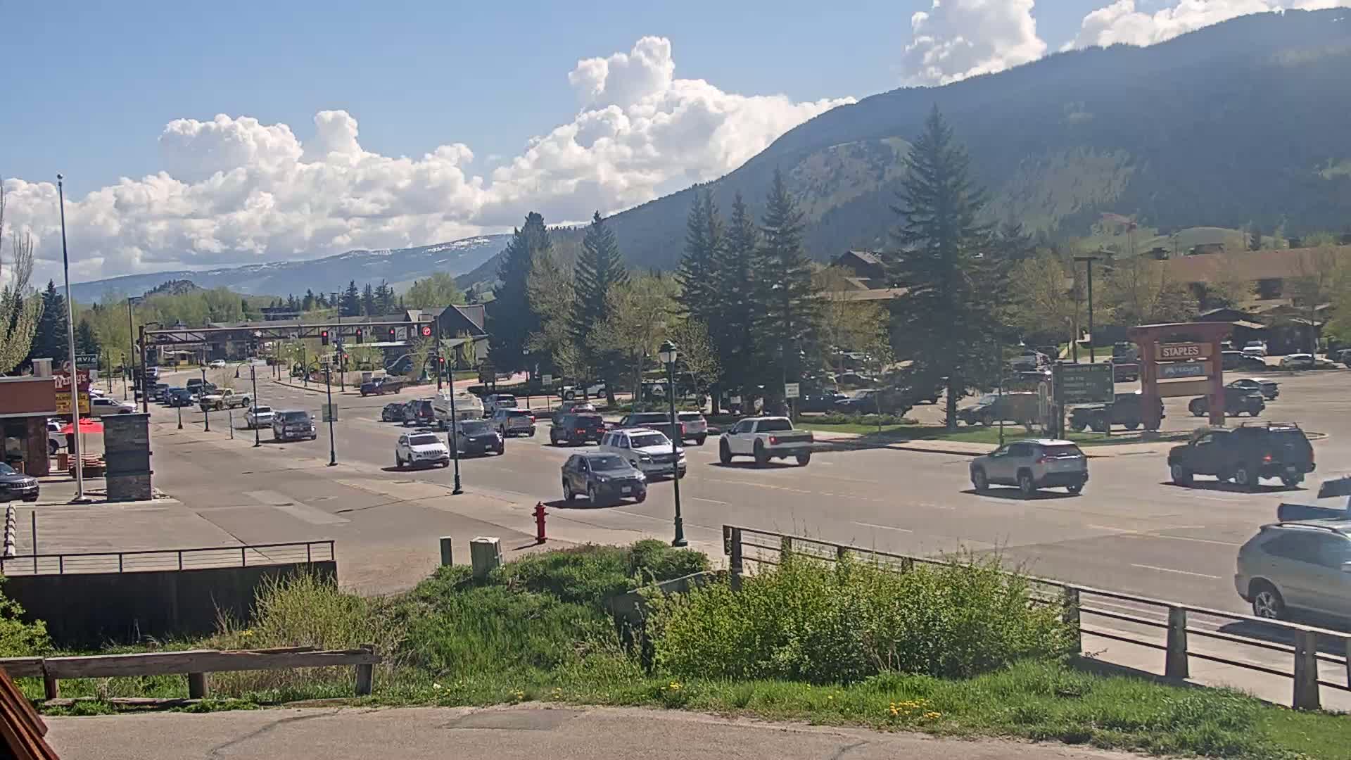 A partly cloudy day reveals a mountain range in the background as cars drive along a multi-lane road through a town with green trees and buildings.