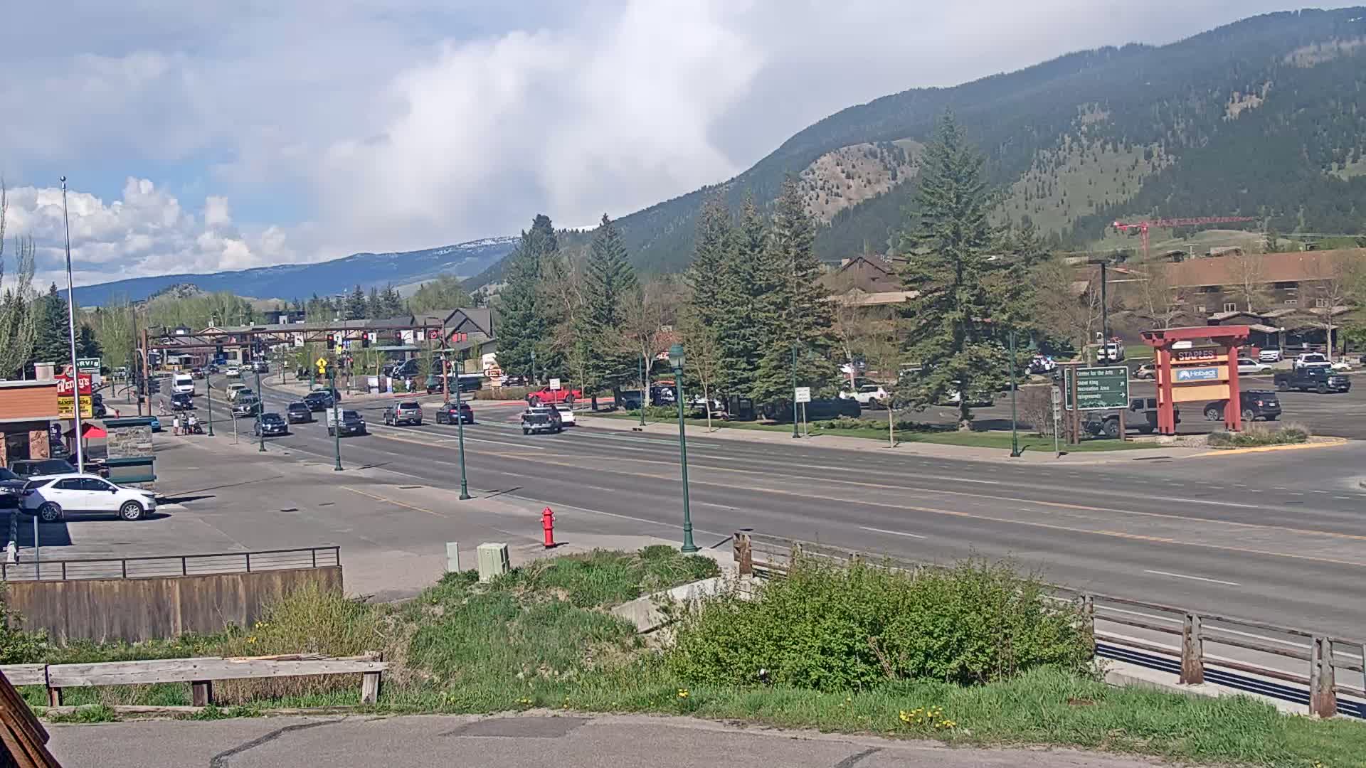 A partly cloudy day reveals a mountain-backed town street scene with several cars driving on a multi-lane road lined with businesses and trees.