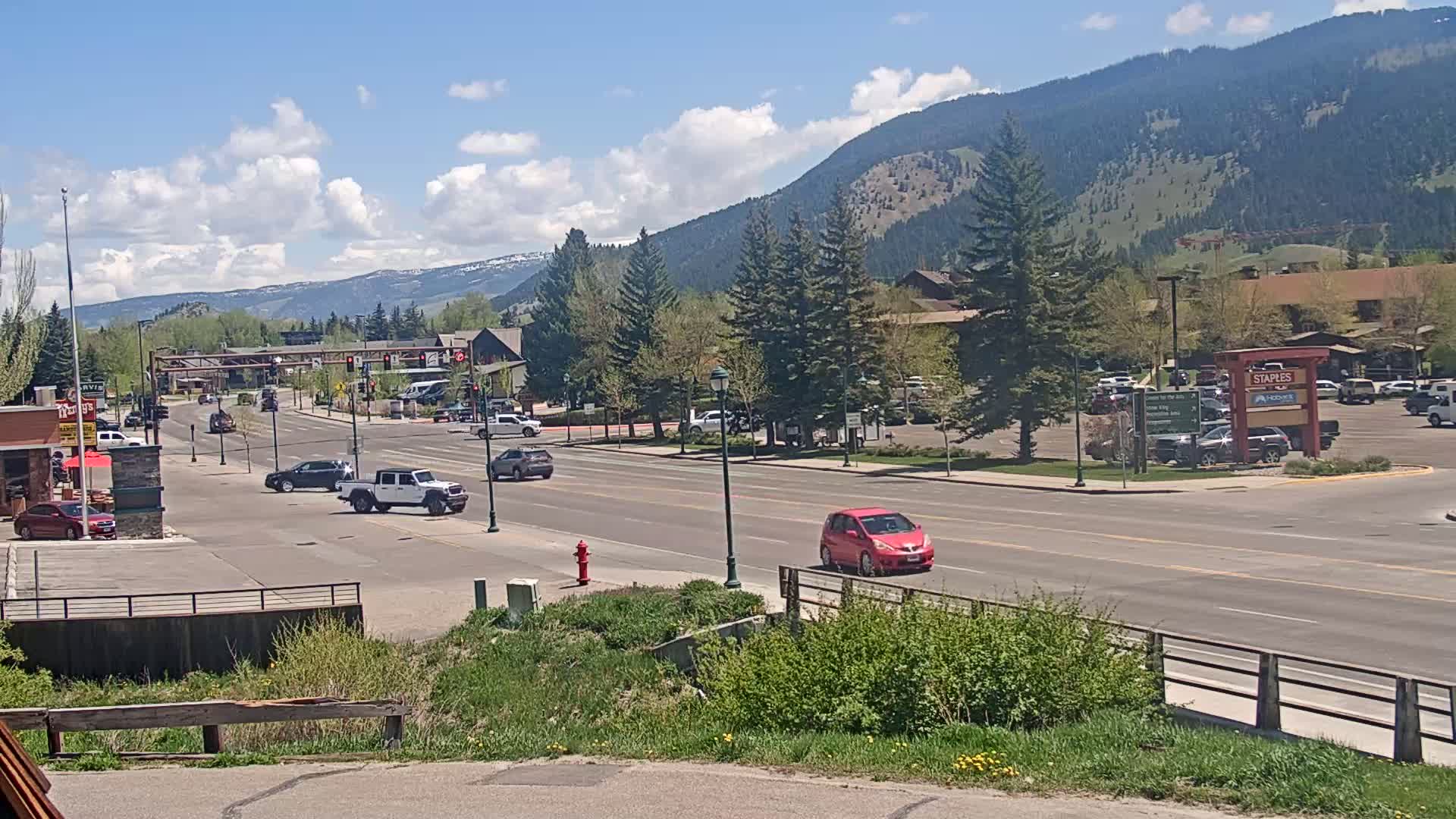 A sunny day reveals a four-lane road in a town nestled in a valley, flanked by buildings, parking lots, and green spaces, with mountains in the background.