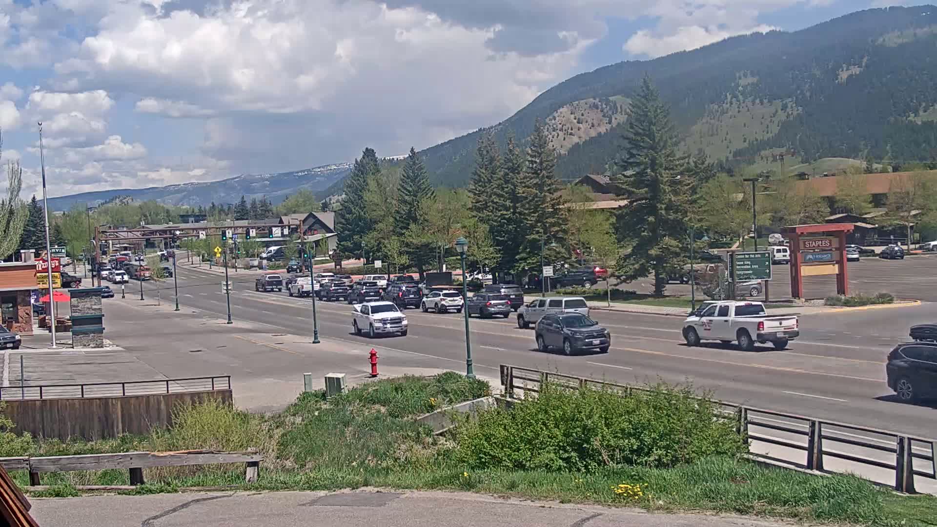 A partly cloudy day with mountains in the background shows a road with moderate traffic, lined with trees and buildings.