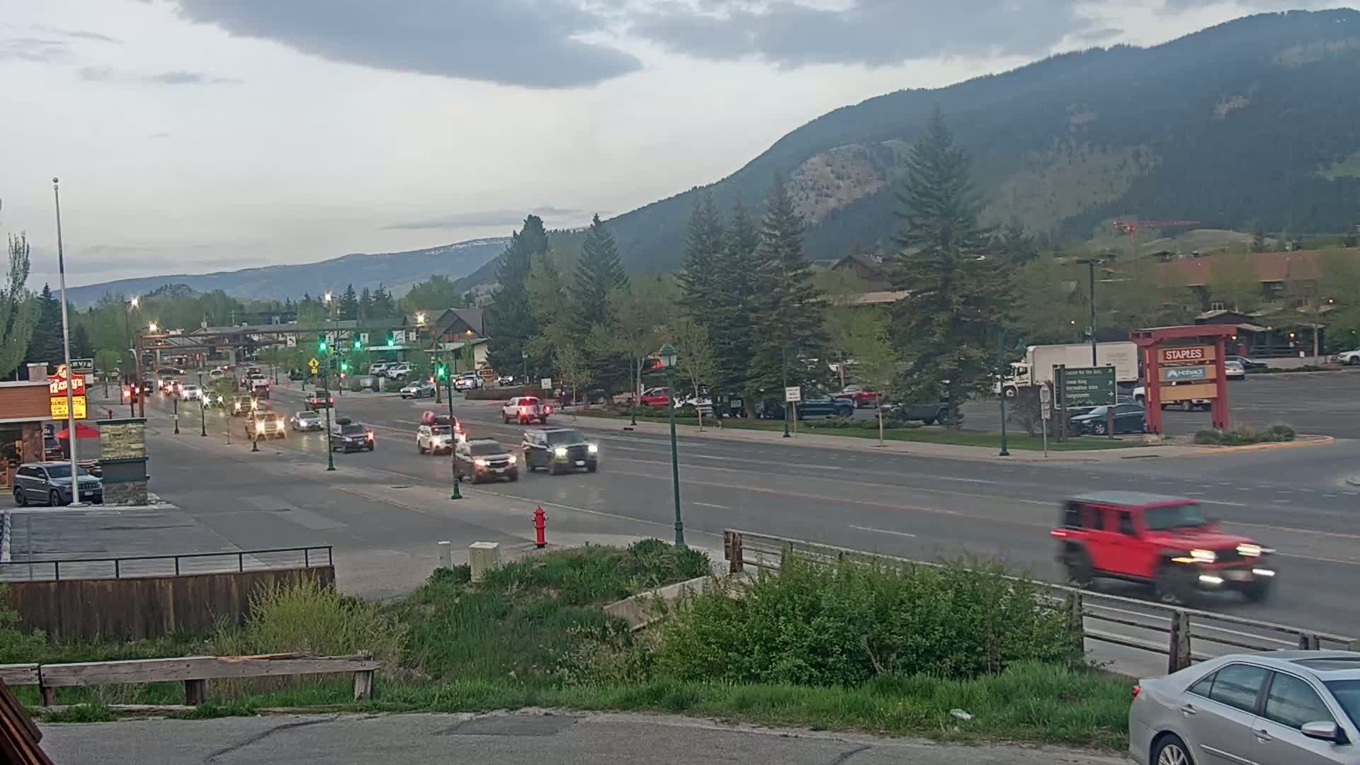 A multi-lane road with moderate traffic flows through a town nestled against a mountain range under an overcast sky.