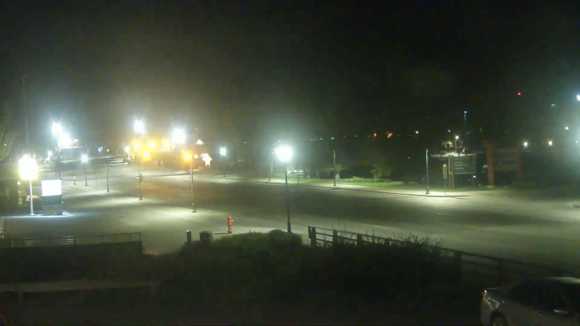 A hazy night scene shows a mostly empty roadway illuminated by streetlights, with a few vehicles visible in the distance and a red fire hydrant near the bottom center.