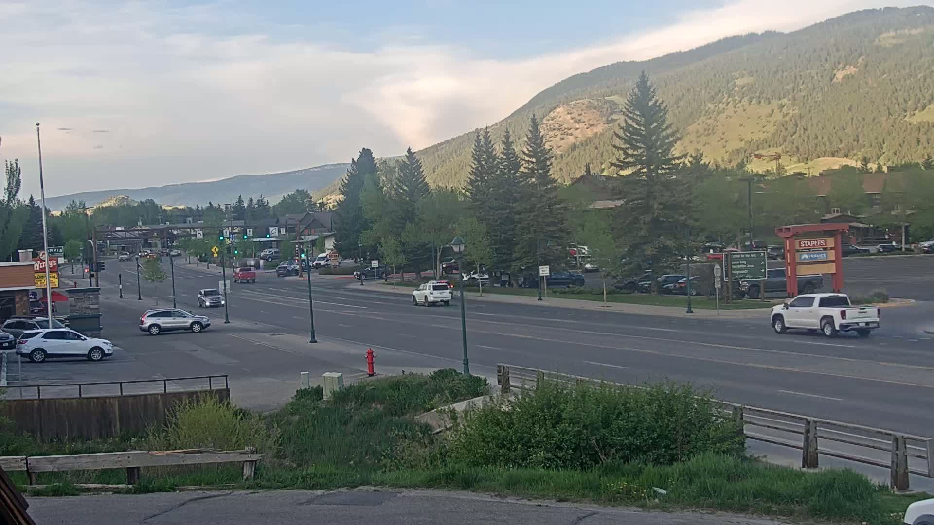 A multi-lane road winds through a town nestled in a valley, under a partly cloudy sky, with mountains rising in the background.