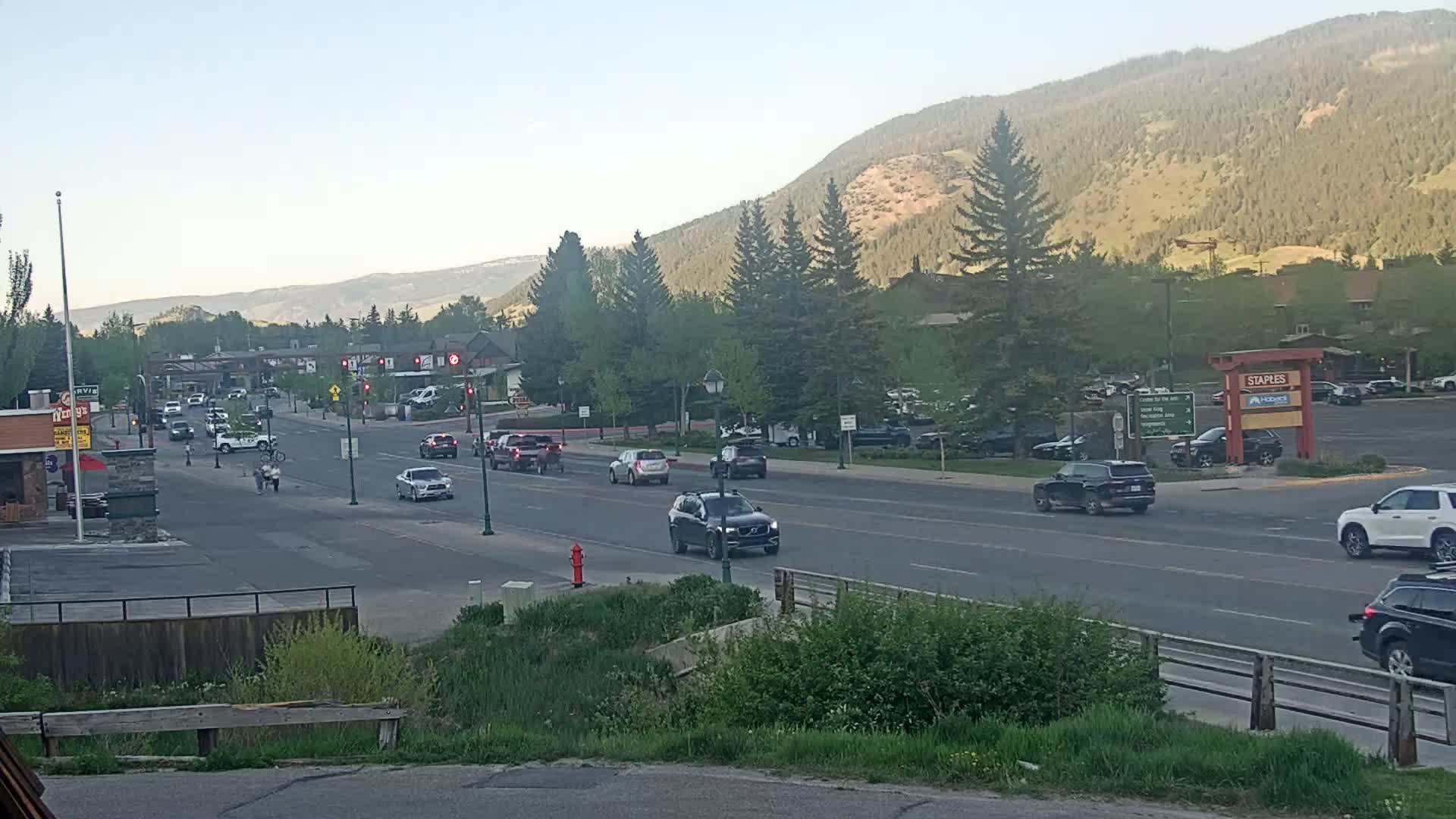 A mostly clear, sunny day reveals a multi-lane road with moderate traffic passing through a town nestled at the base of a mountain range.