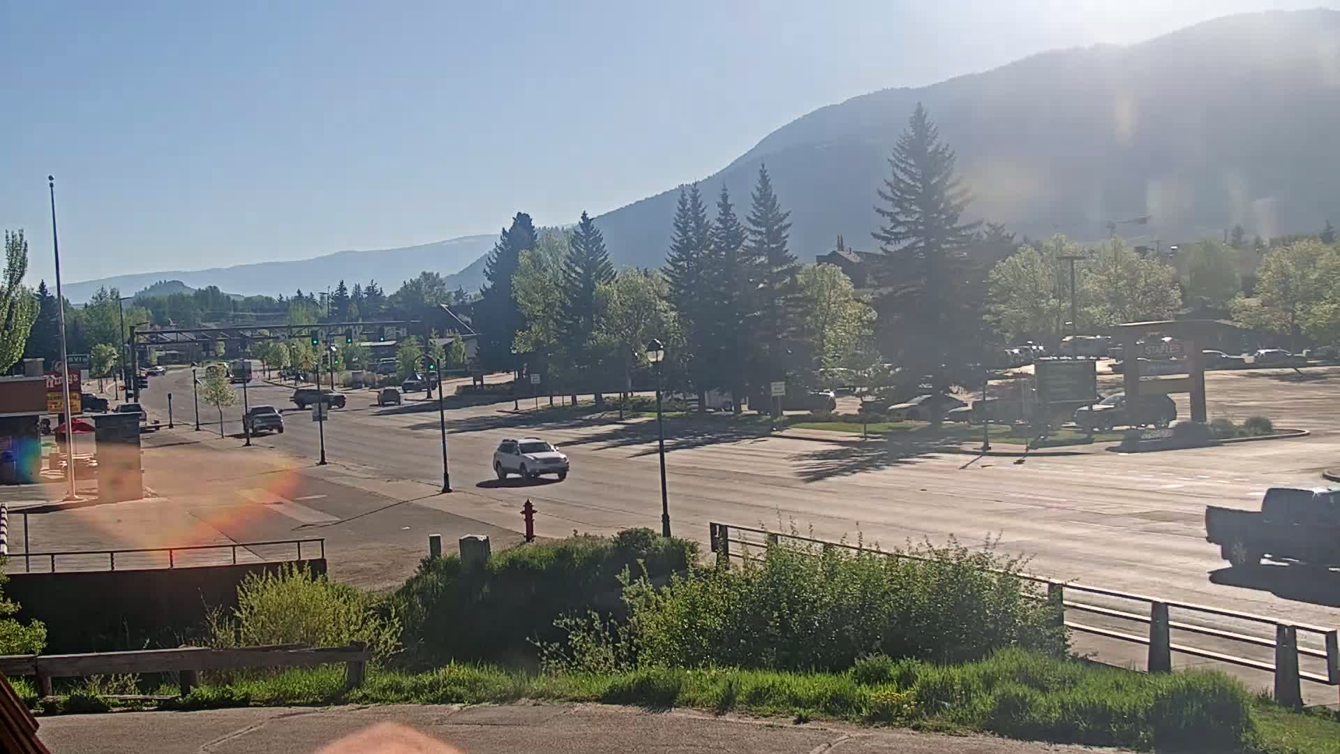 A mostly sunny day shows a road with sparse traffic passing through a town nestled at the base of hazy mountains, lined with trees and some buildings.