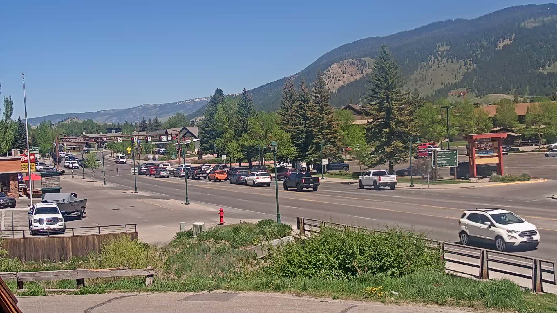 A sunny day reveals a road lined with cars, leading toward a mountain range and buildings nestled among green trees.