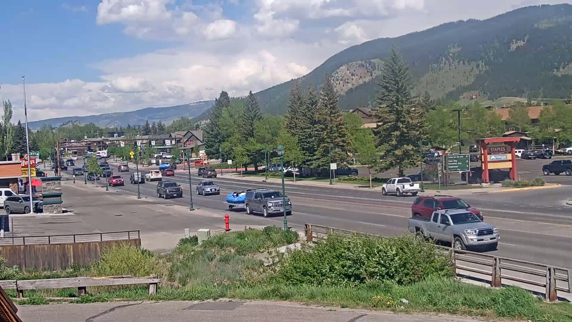 A partly sunny day shows a main road with moderate traffic passing through a town nestled in a valley surrounded by mountains.