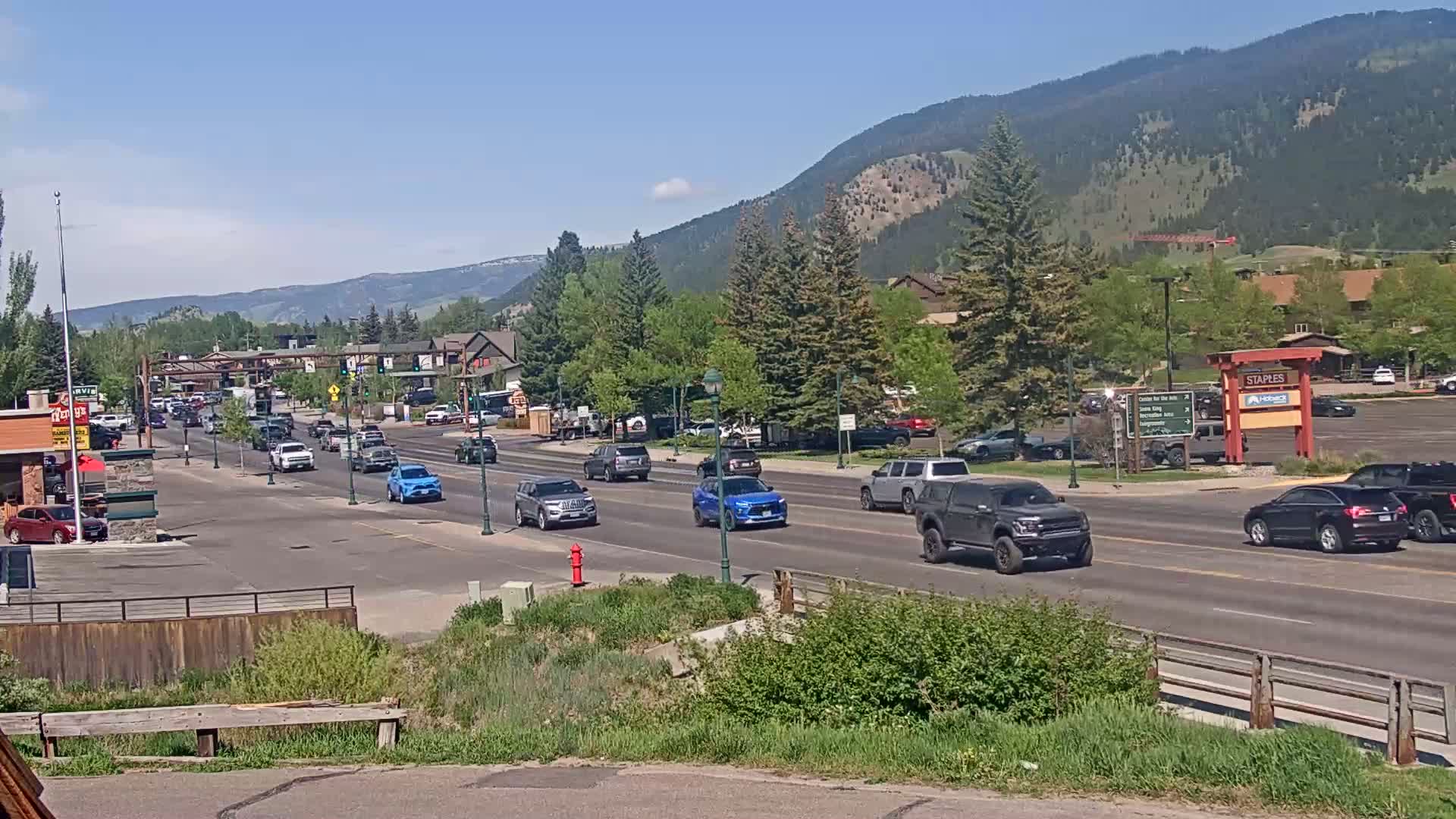 A sunny day reveals a mountain view behind a town's main street with several cars driving along it.