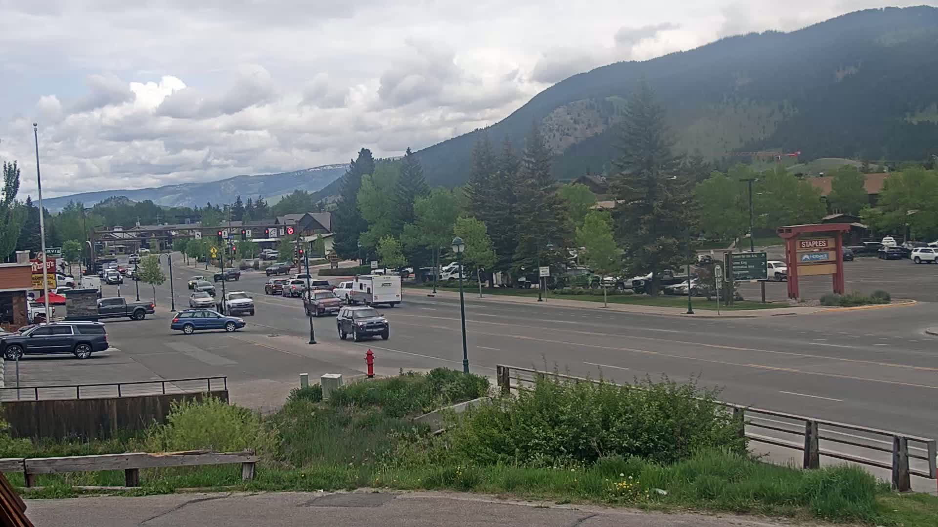 A partly cloudy day shows a mountain-backed town with cars driving down a road lined with trees and buildings.