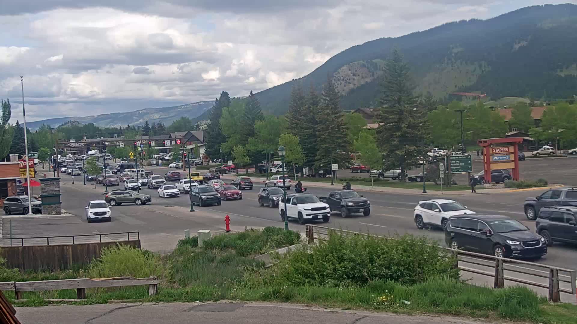 A busy street scene in a mountainous town with overcast skies shows numerous cars driving through an intersection with lush green trees and buildings along the sides of the road.