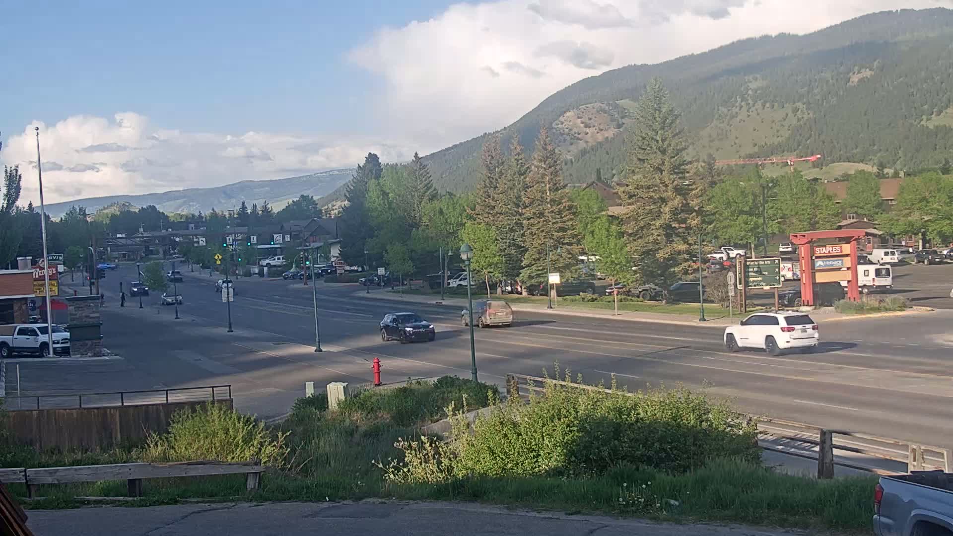 A partly cloudy day reveals a mountain town street scene with several cars driving on a multi-lane road, bordered by businesses, trees, and a parking lot.