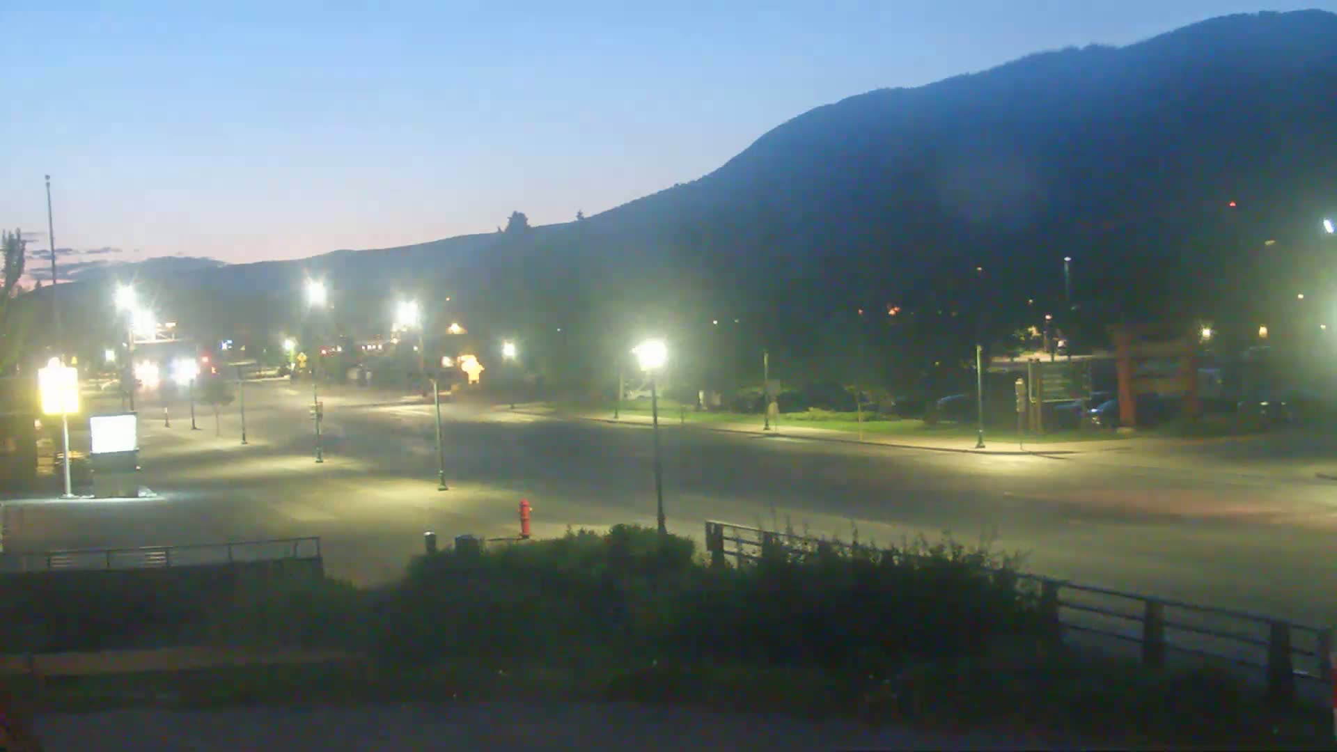 An empty, well-lit street at twilight is situated below a dark mountain range under a clear, dusky sky.