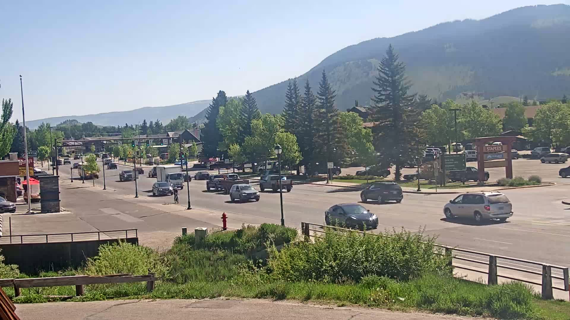 A sunny day reveals a town's main street with moderate traffic, lined with buildings and trees, nestled against a backdrop of hazy mountains.