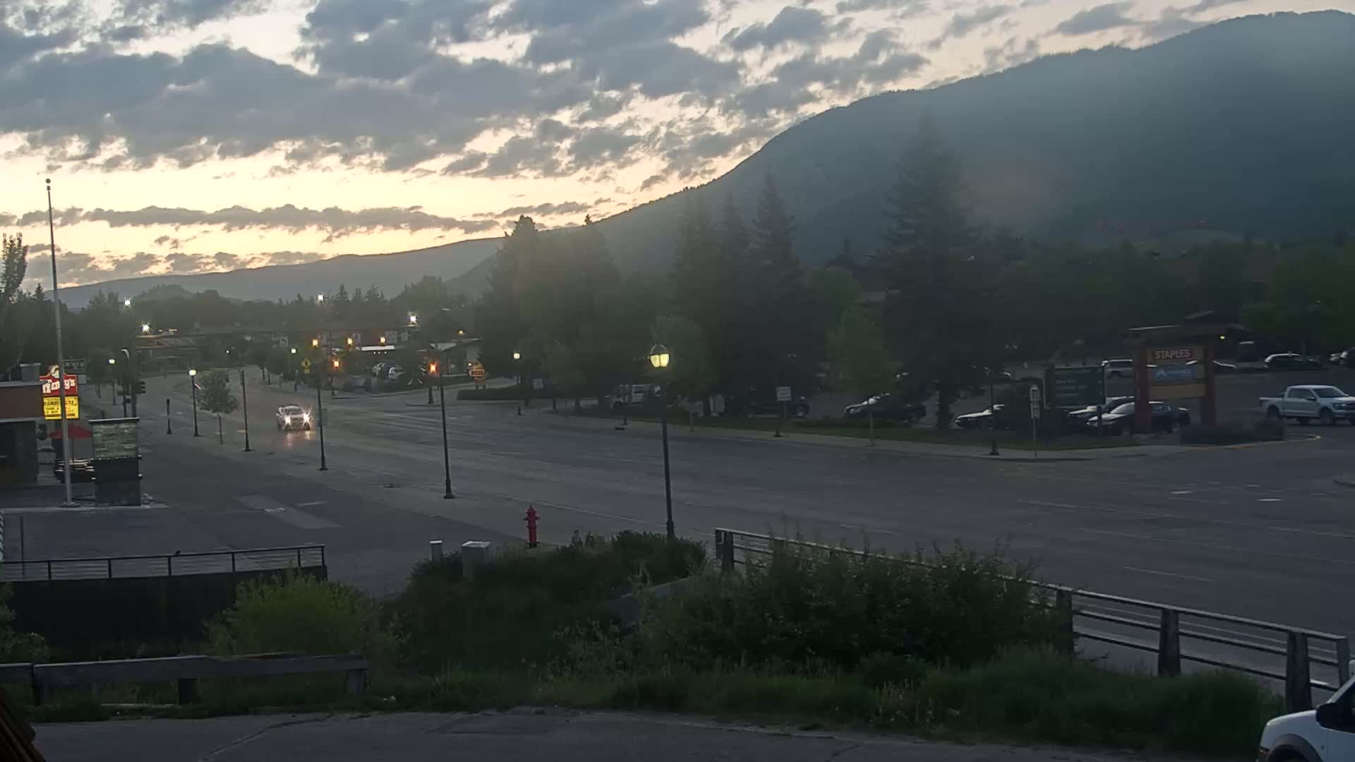 A mostly empty street with a few cars under a cloudy sunrise, with mountains in the background.