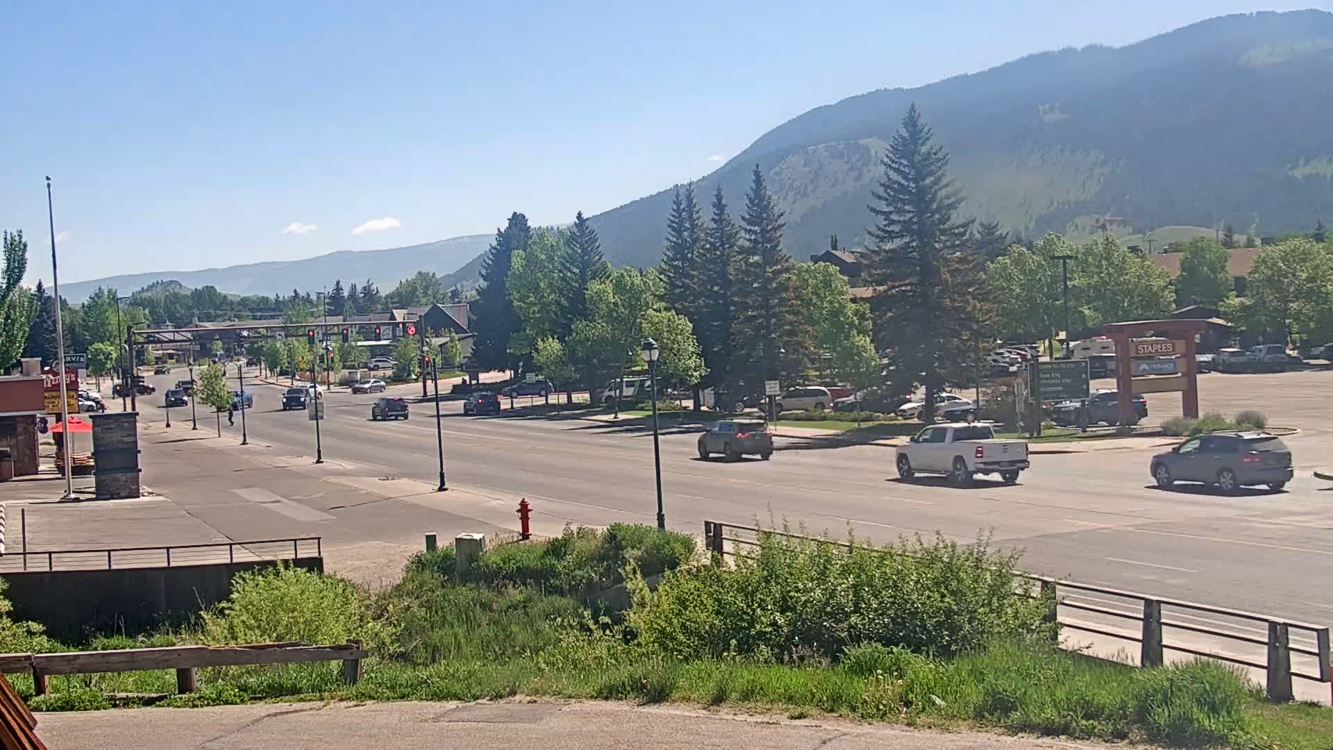 A sunny day reveals a multi-lane road with moderate traffic, lined with green trees and buildings, set against a backdrop of distant mountains.