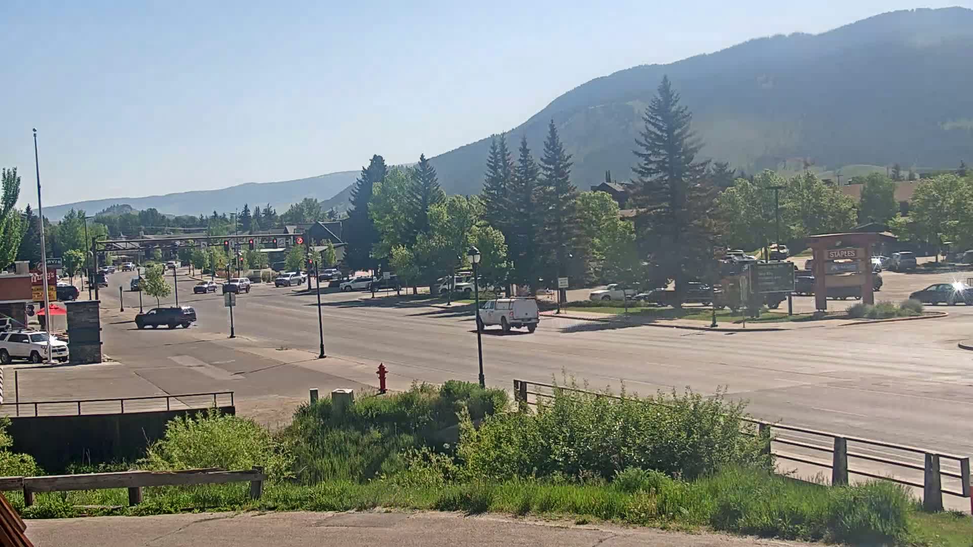 A mostly sunny day reveals a town's main street with light traffic, lined with trees and buildings, set against a backdrop of hazy mountains.