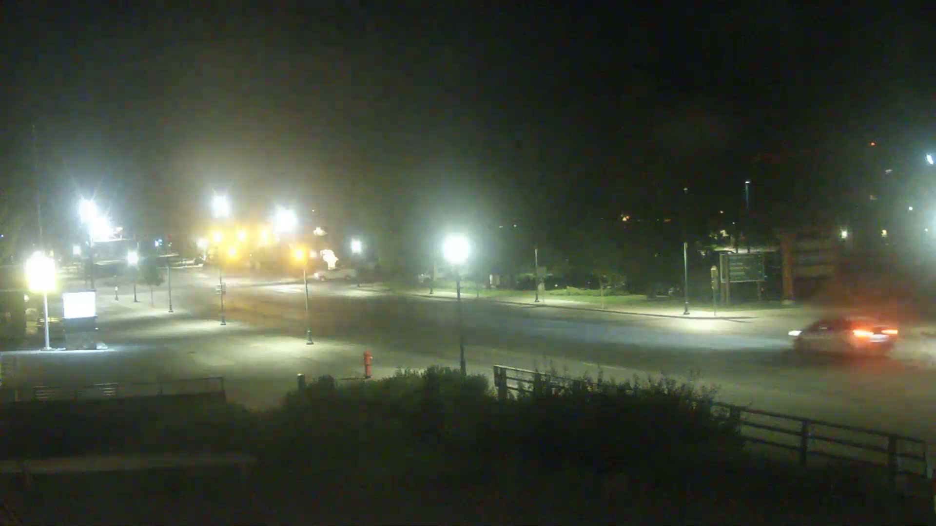 A hazy night scene shows a mostly empty road intersection with several streetlights illuminating the area and a single car driving on the road.
