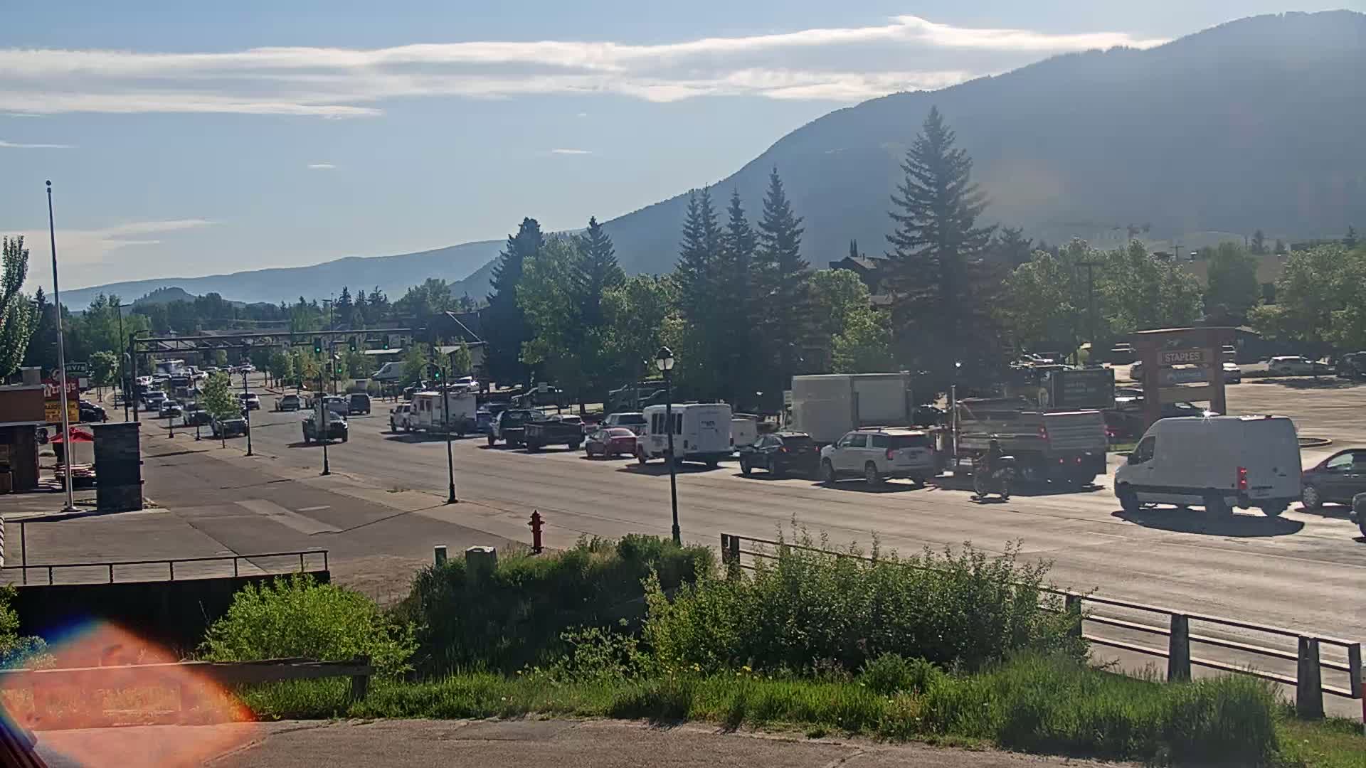 A sunny day reveals a line of vehicles stopped in traffic on a road bordered by green vegetation, with mountains visible in the background.