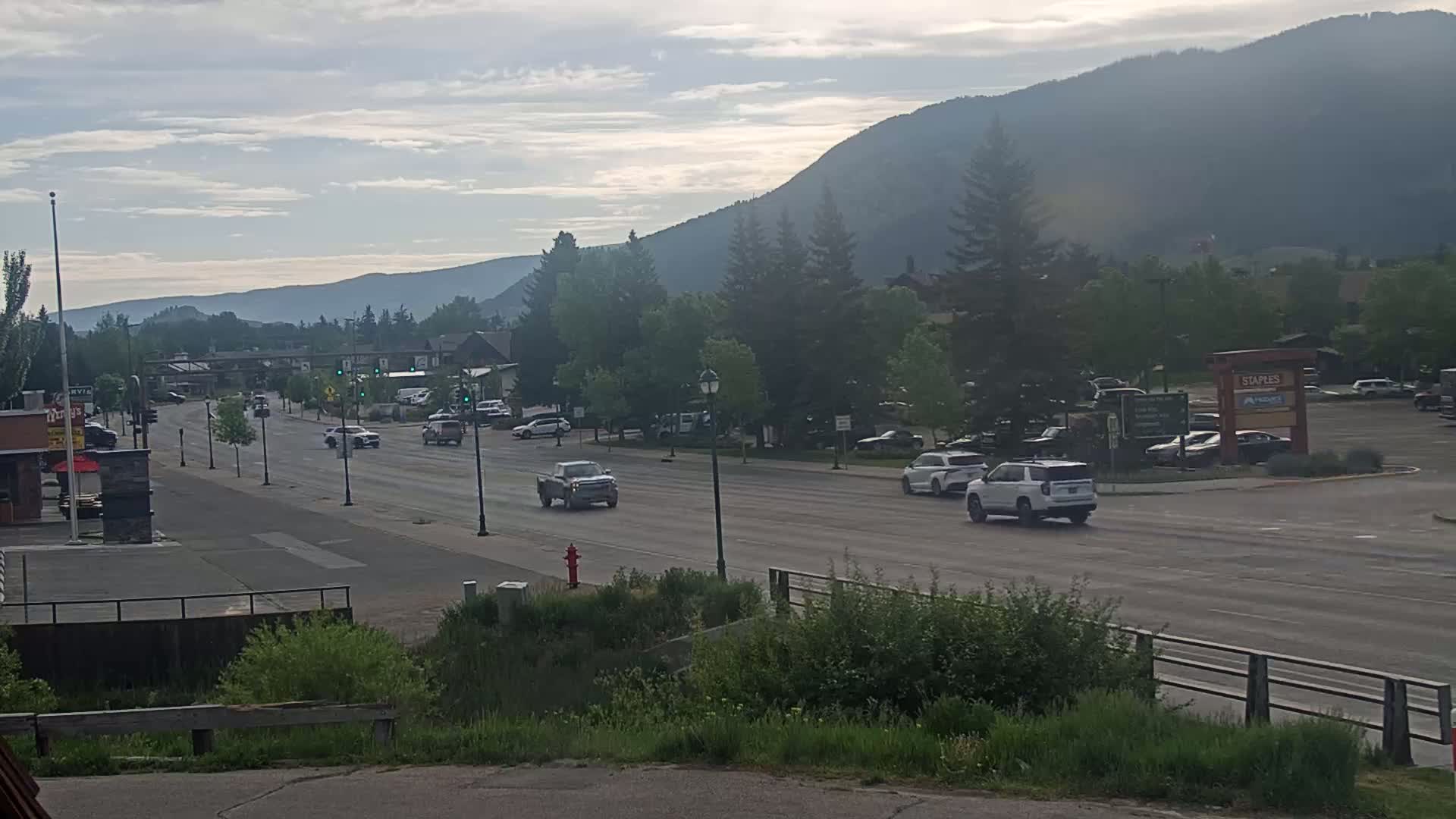 A mostly clear, slightly hazy day reveals a mountain range in the background, overlooking a paved road with several vehicles and lush green vegetation on either side.