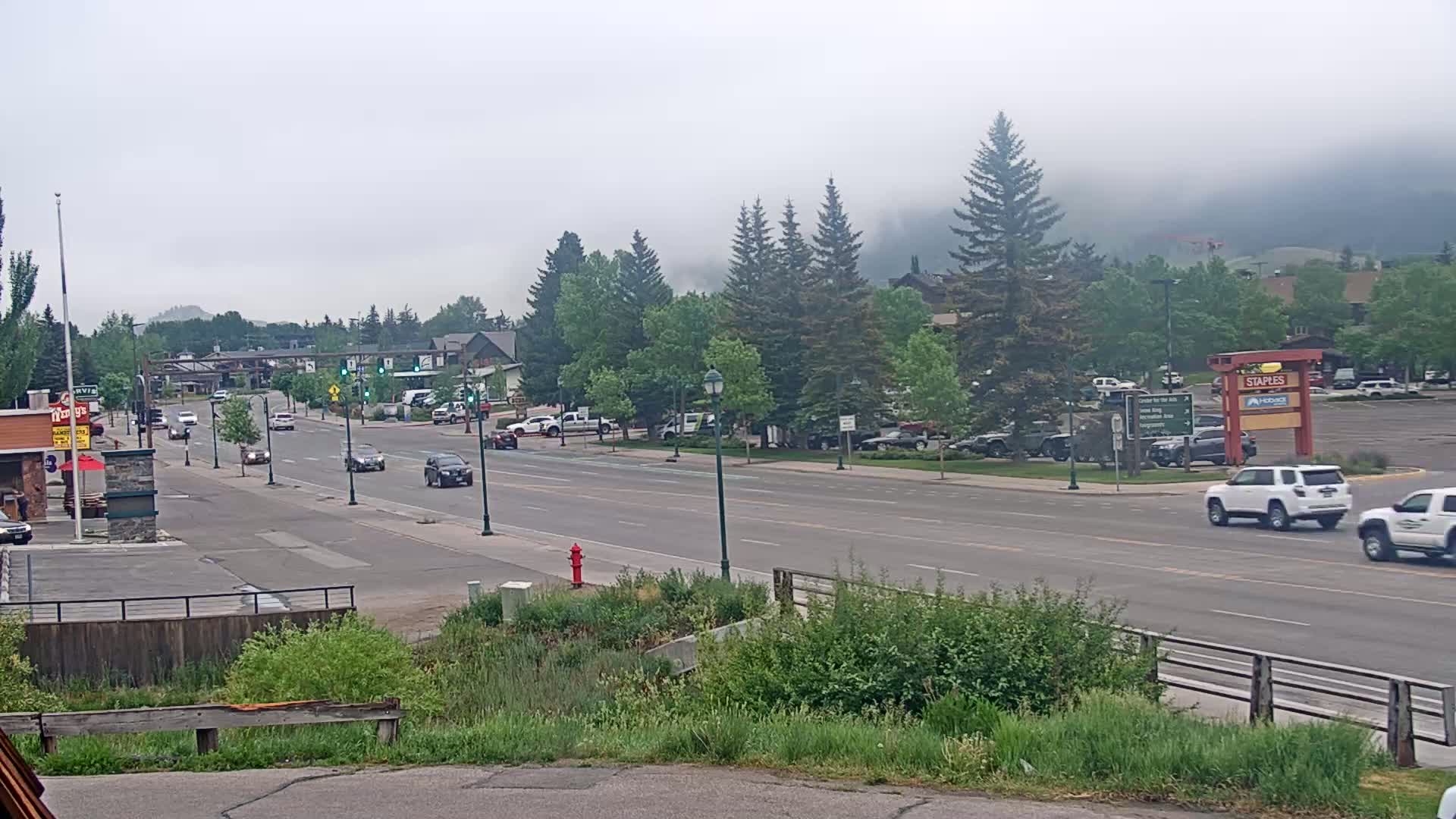A multi-lane road with light traffic passes through a town, lined with trees and buildings, under a hazy sky.