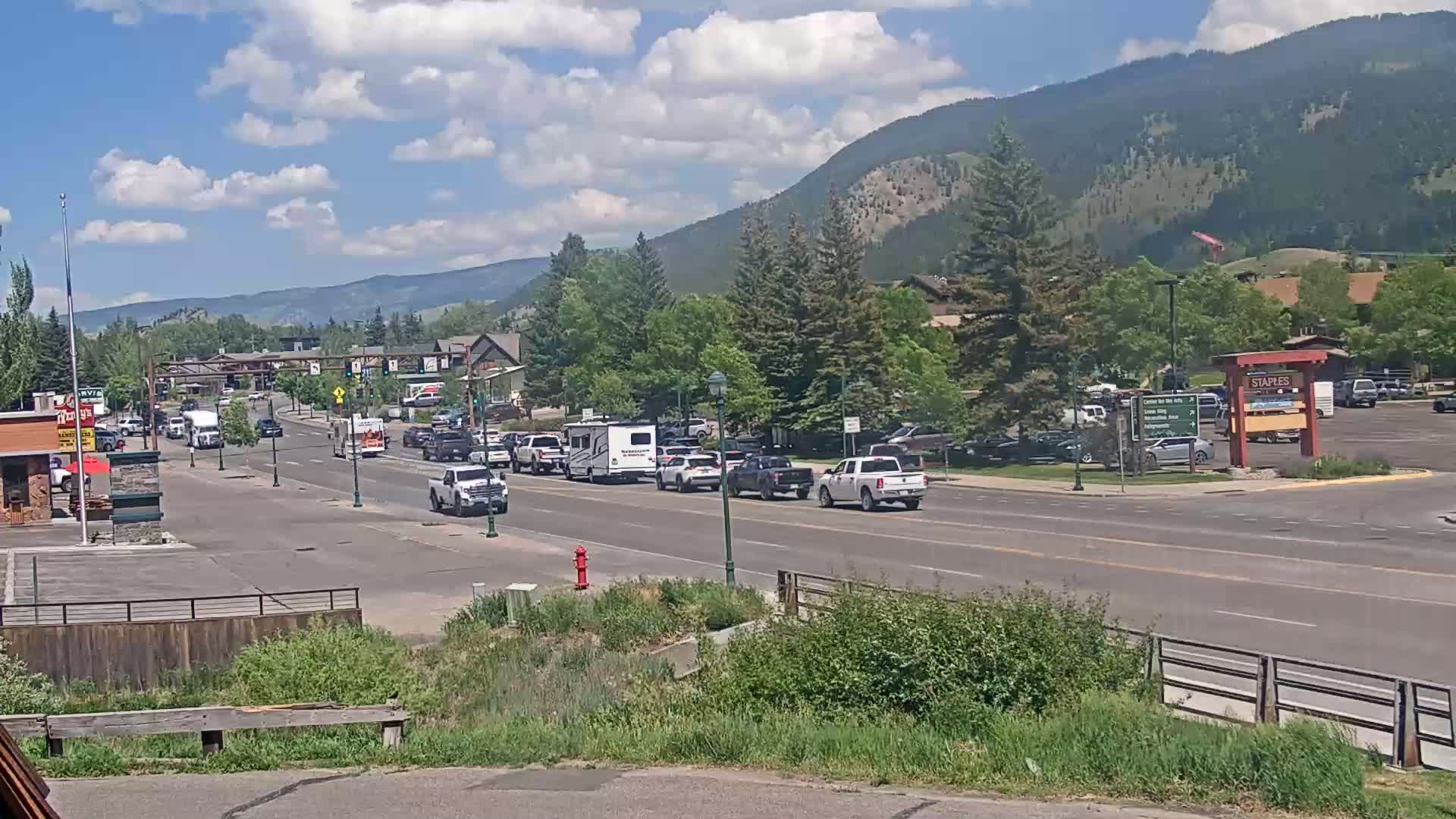 A partly cloudy day shows a street scene with moderate traffic, including several RVs, in a town nestled at the base of a mountain range.