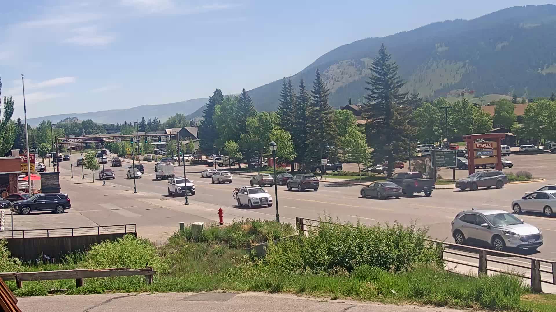 A sunny day with clear skies shows a multi-lane road with moderate traffic passing through a town, set against a backdrop of mountains and green trees.
