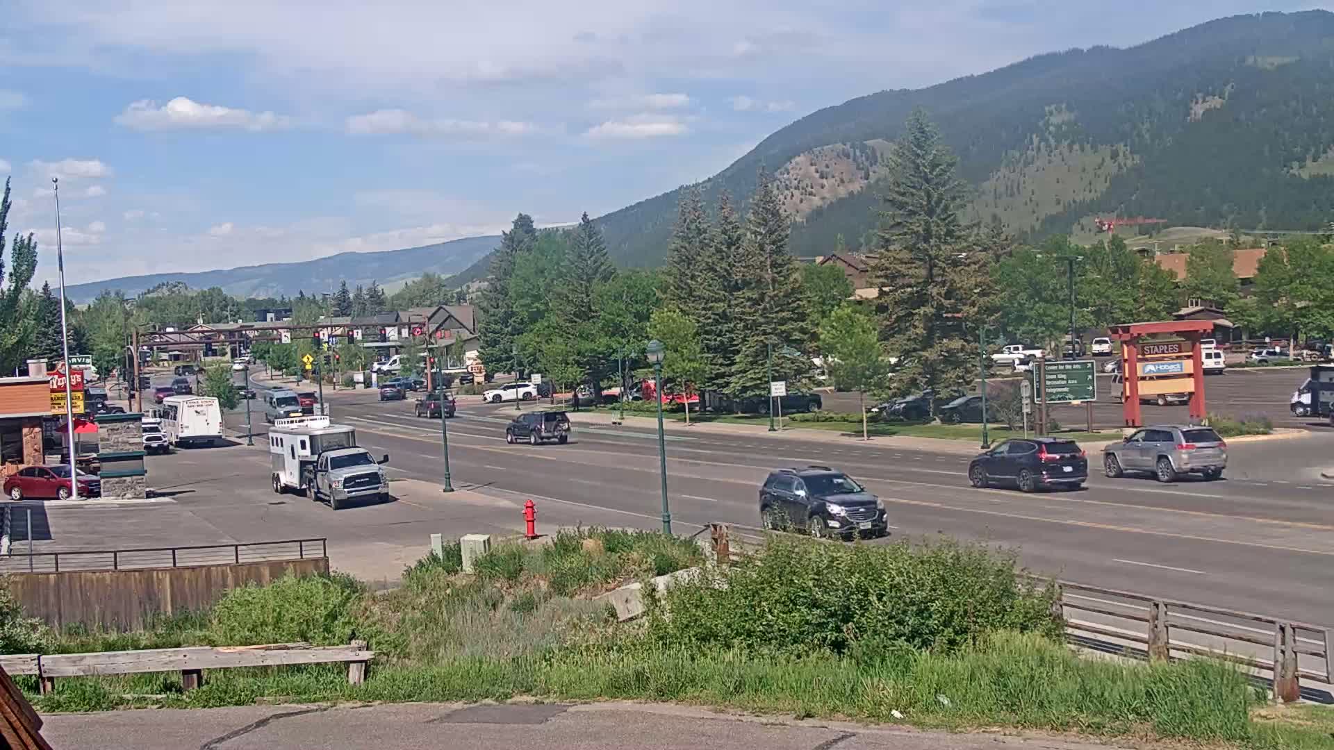 A sunny day with clear skies shows a multi-lane road with moderate traffic passing through a town, set against a backdrop of mountains and green trees.