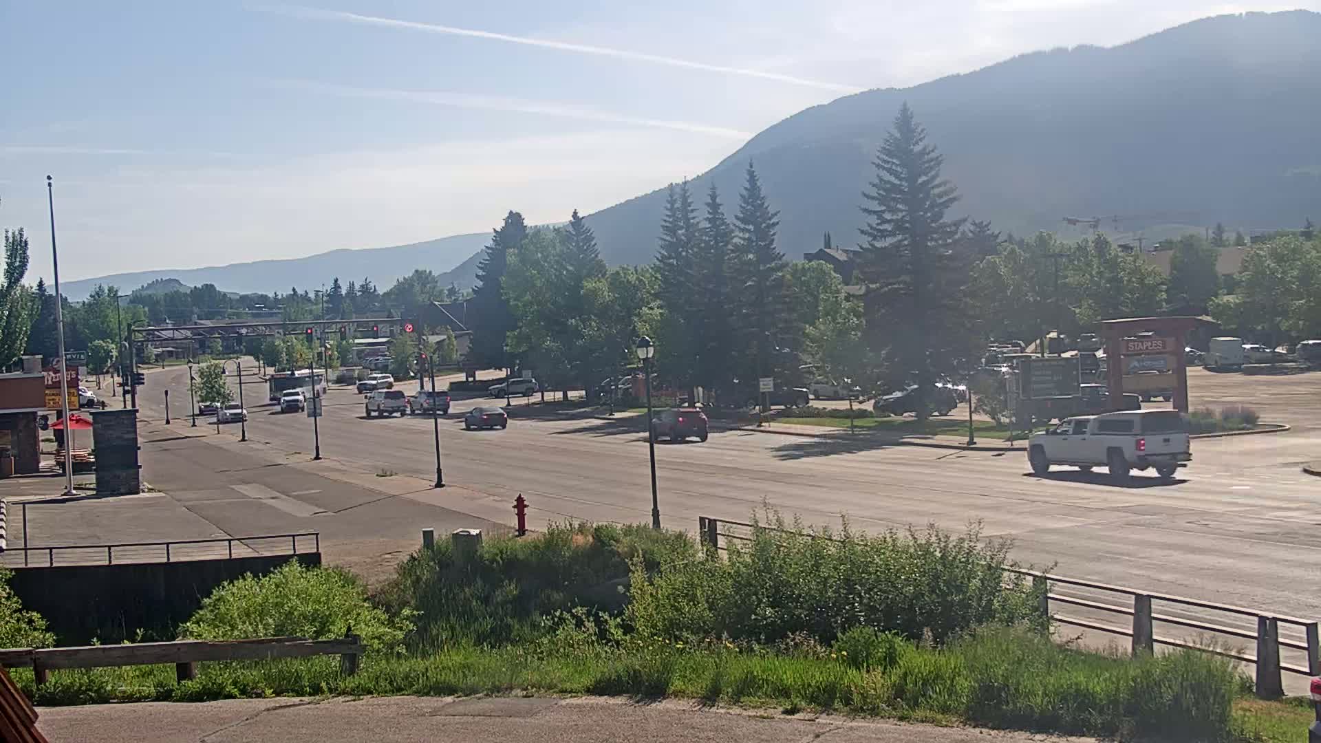A sunny day reveals a multi-lane road with sparse traffic, lined with green vegetation and buildings, situated against a backdrop of hazy mountains.