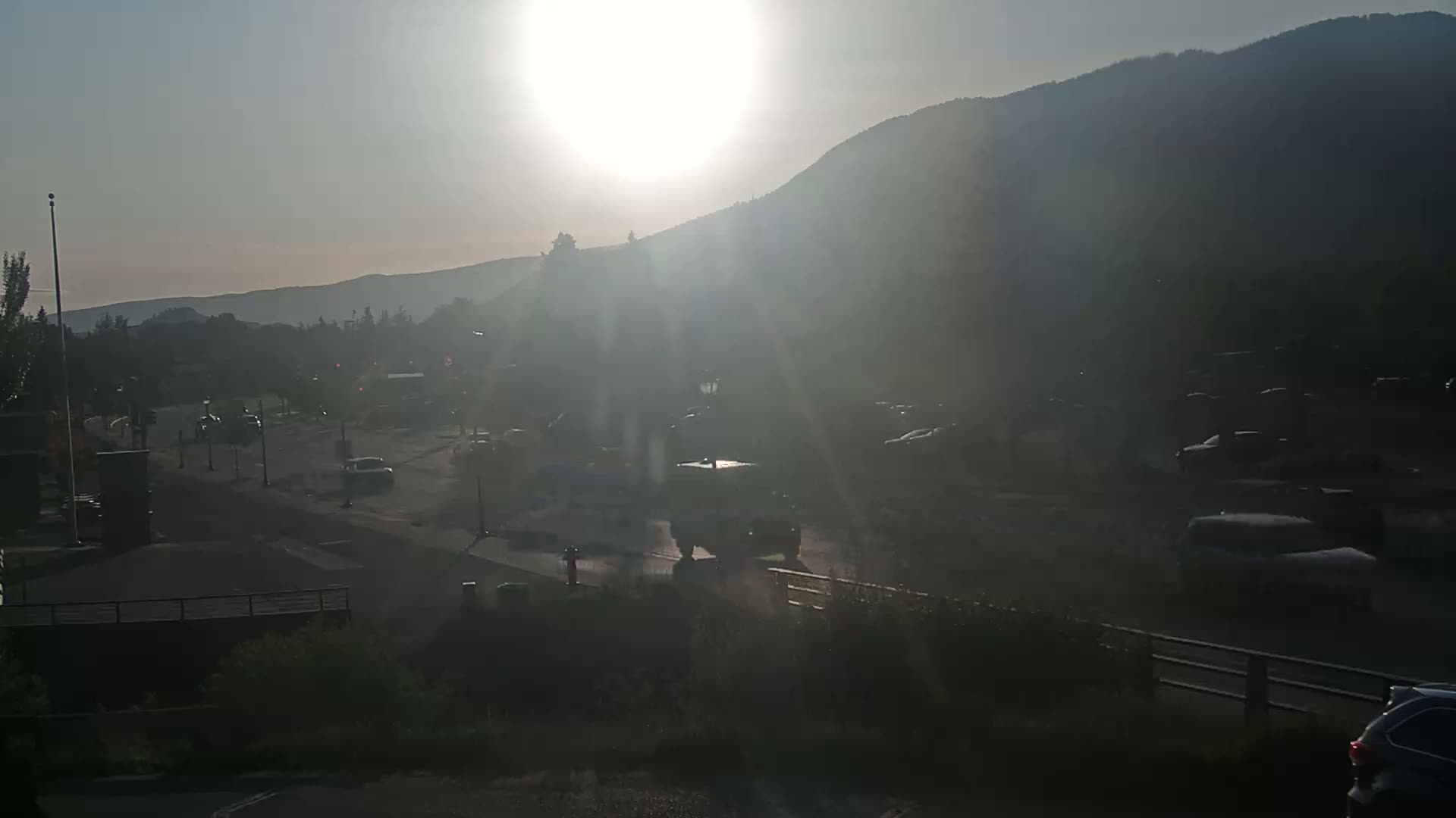 A hazy sunrise illuminates a mountain range behind a street with several vehicles, creating bright sun rays.