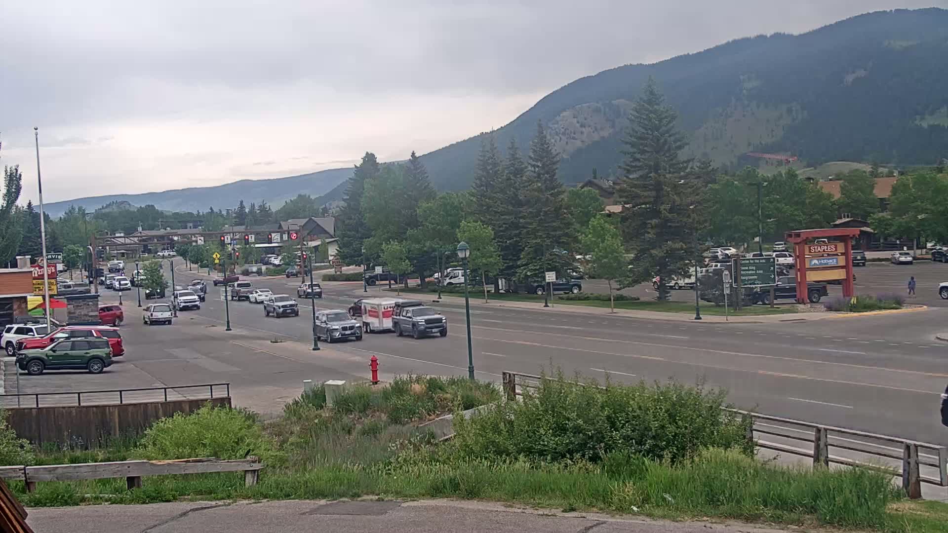 A partly cloudy day reveals a street scene with moderate traffic, lined with buildings and trees, set against a backdrop of mountains.