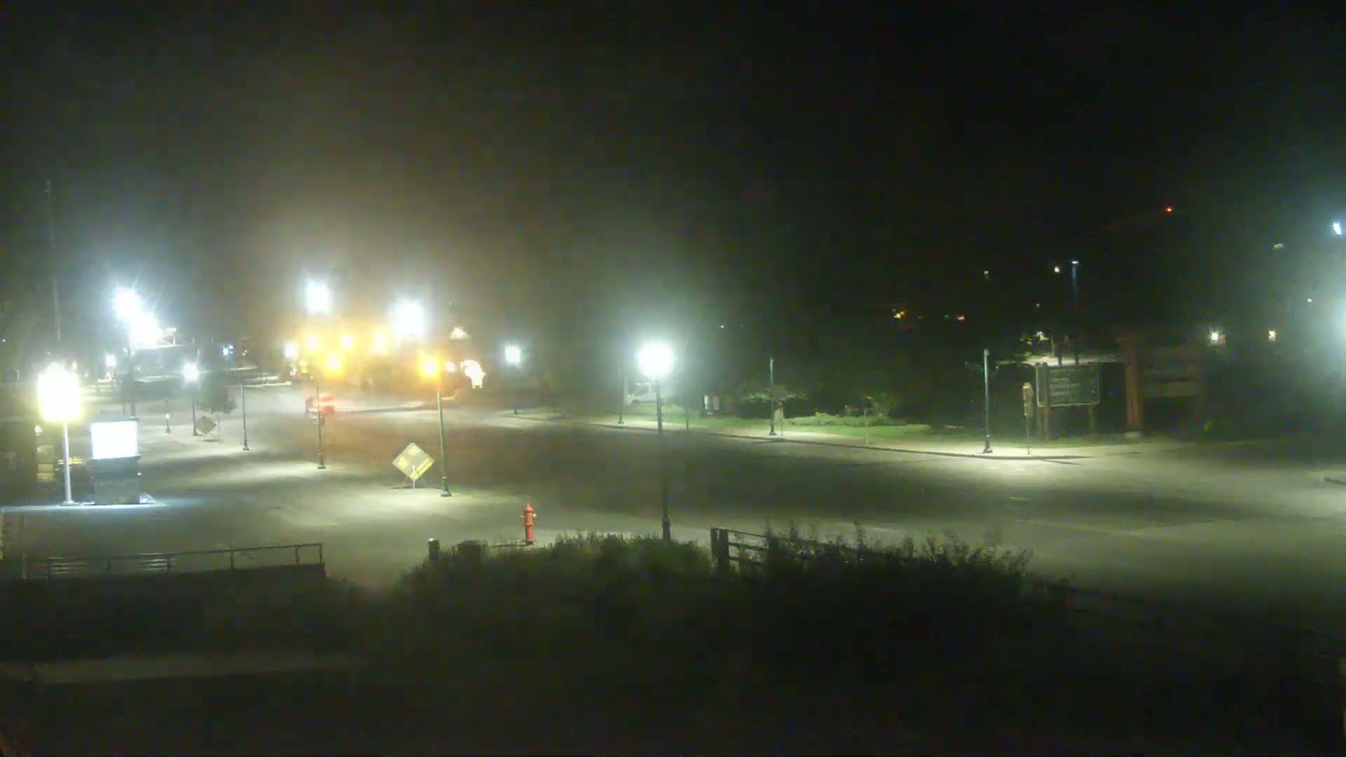 A foggy night scene shows a sparsely trafficked intersection with streetlights illuminating a roadway and surrounding vegetation.