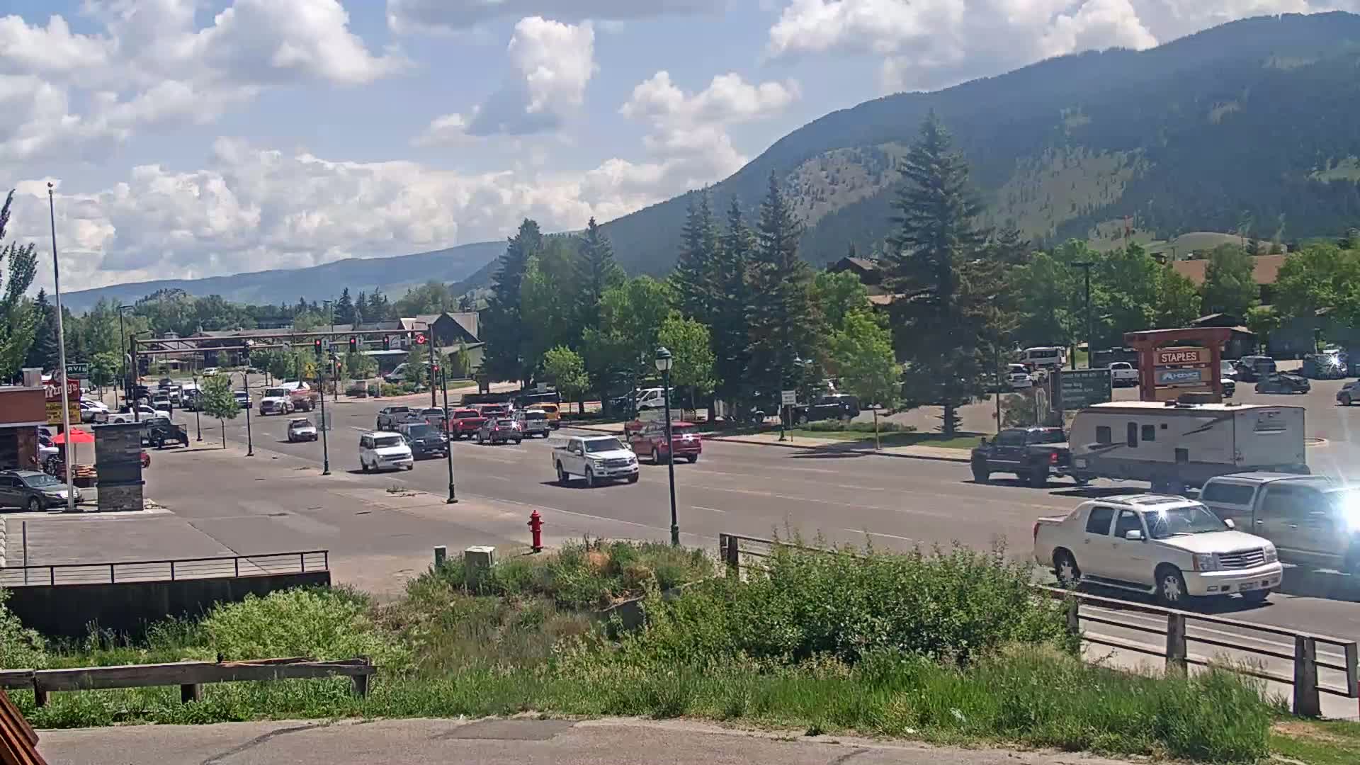 A partly cloudy day shows a mountain range in the background, with moderate traffic on a street lined with trees and buildings.
