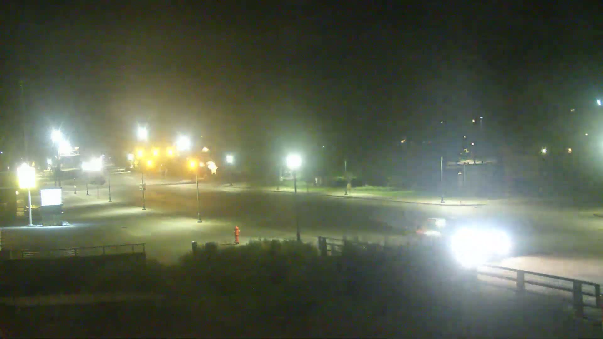 A hazy night scene shows a mostly empty parking lot and roadway with several streetlights illuminating the area, and a few vehicles are visible in the distance.